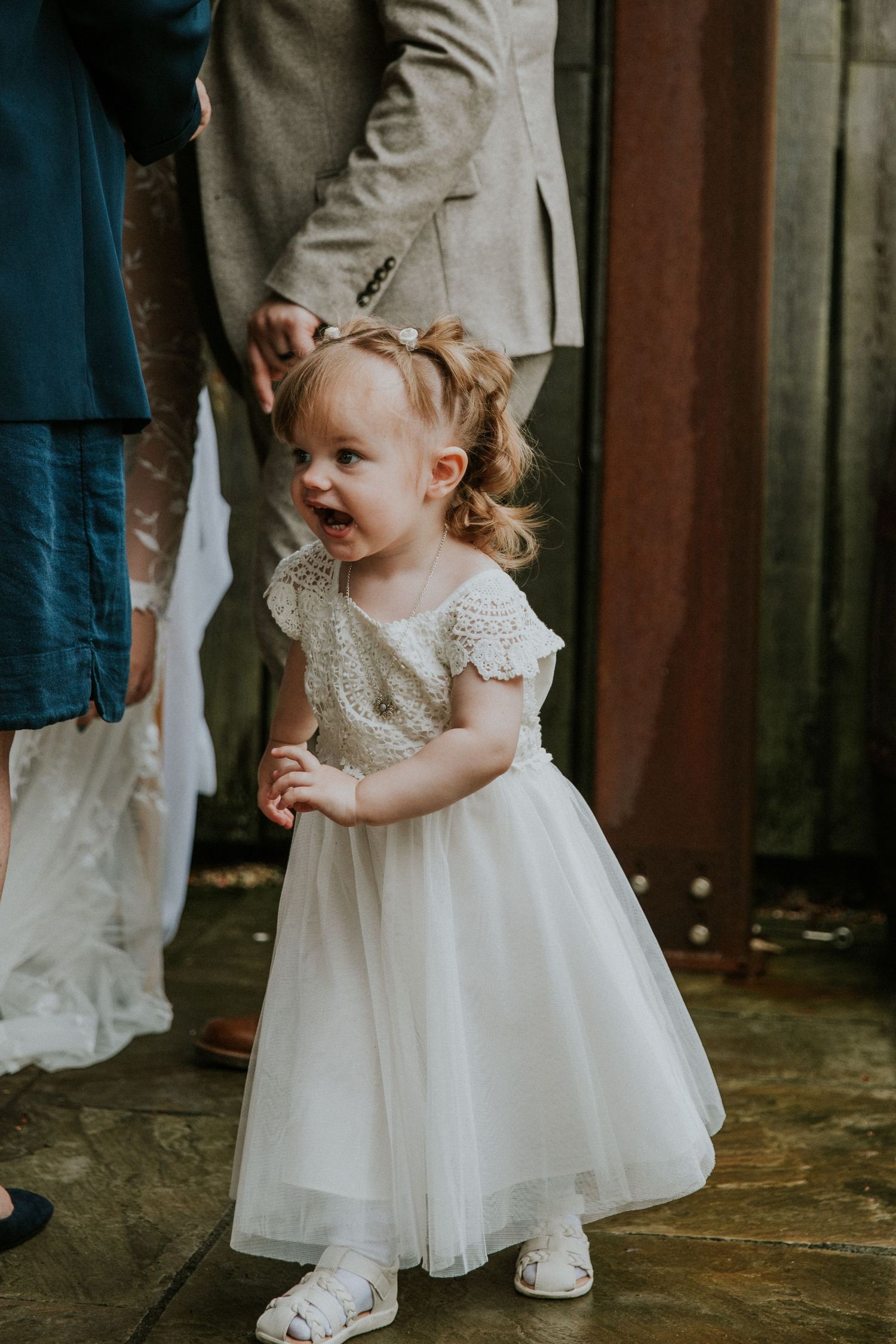 Young child wearing a white lace dress smiles joyfully during a wedding celebration.