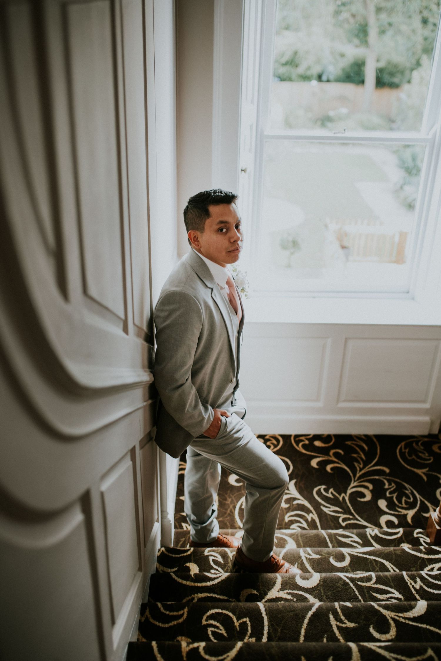 A person in a light gray suit stands thoughtfully by a bright window with ornate carpeting.