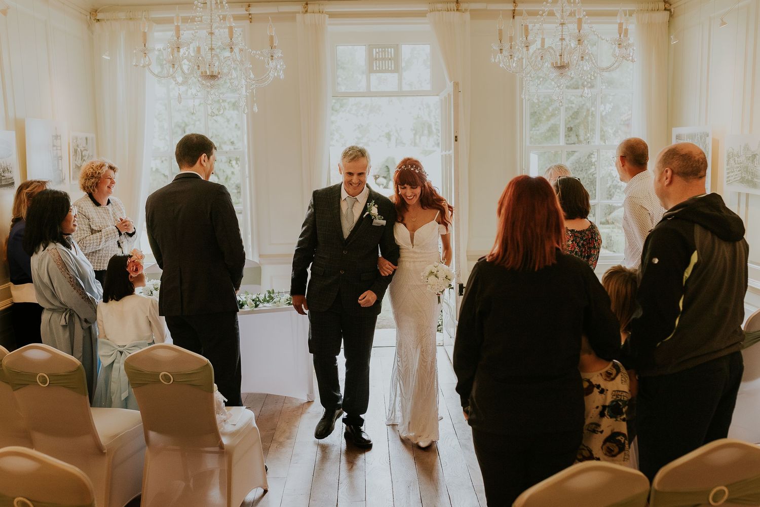 Wedding guests gather in bright ceremony room with large windows and white walls.