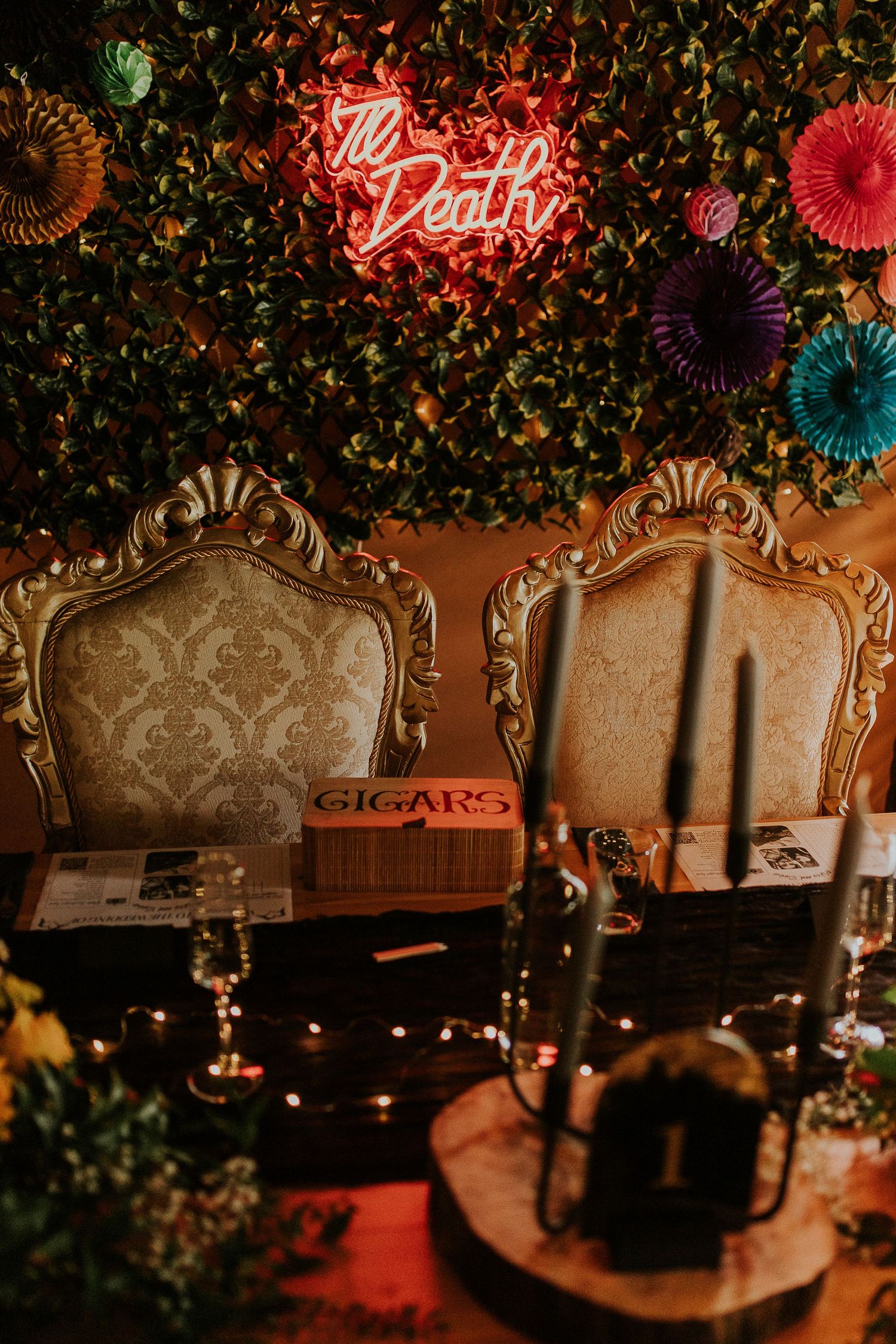 Vintage ornate chairs and cigar box under neon 'Till Death' sign with foliage wall decor and string lights.