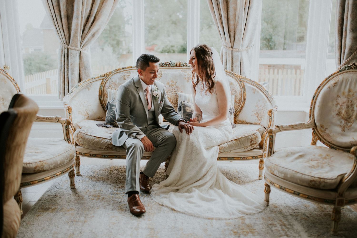 Bride and groom share intimate moment on vintage sofa in sunlit room.