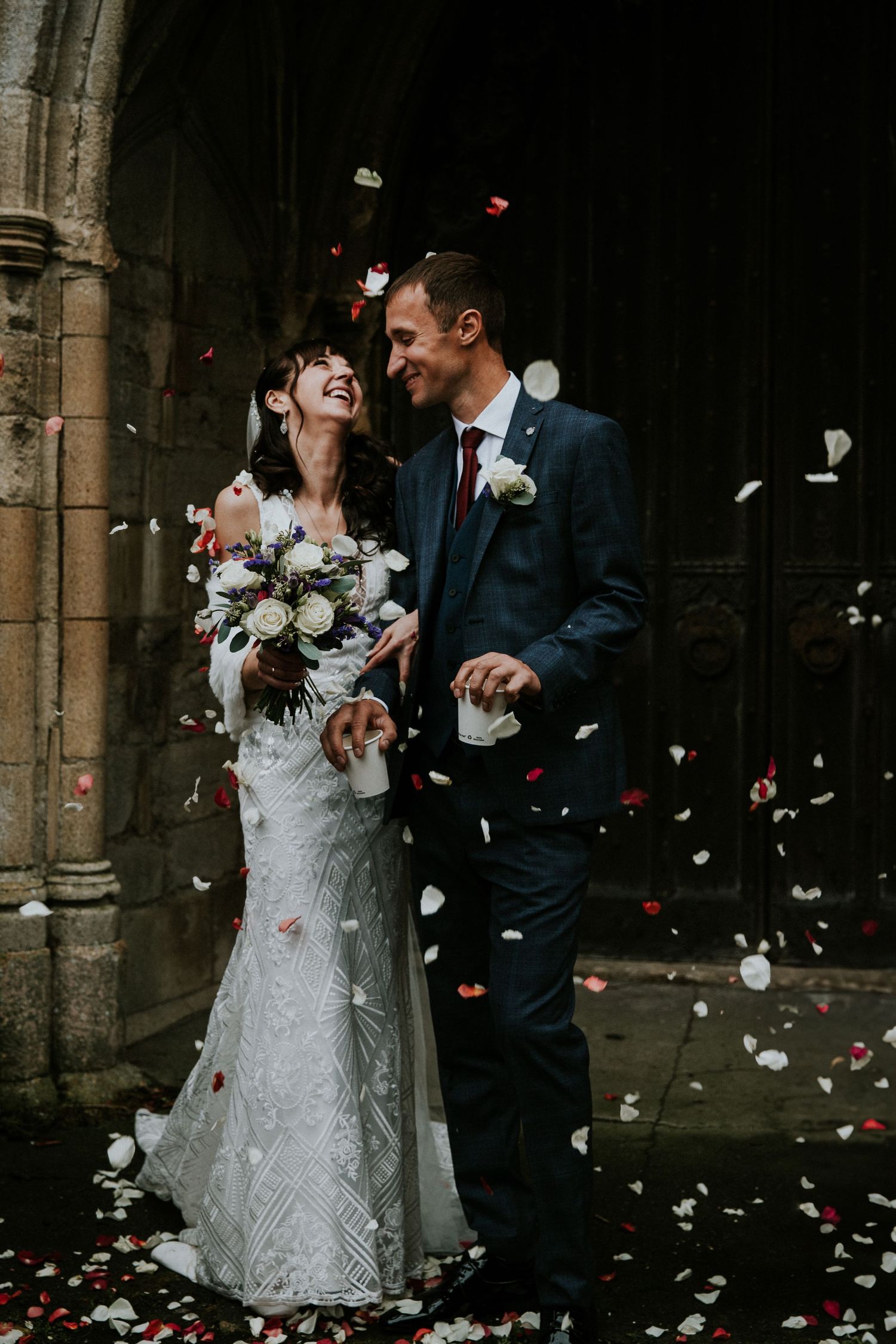 Wedding couple shares joyful moment as rose petals fall around them in church doorway.