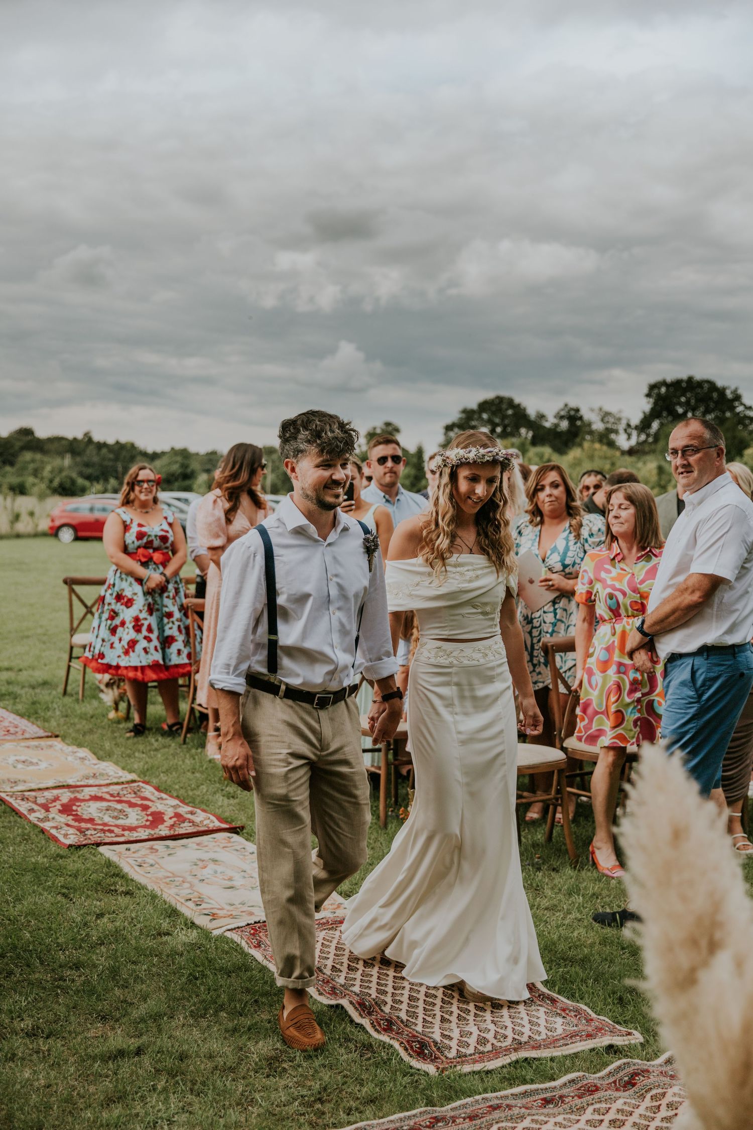 Couple walks down outdoor aisle lined with vintage rugs during boho wedding ceremony under cloudy sky.