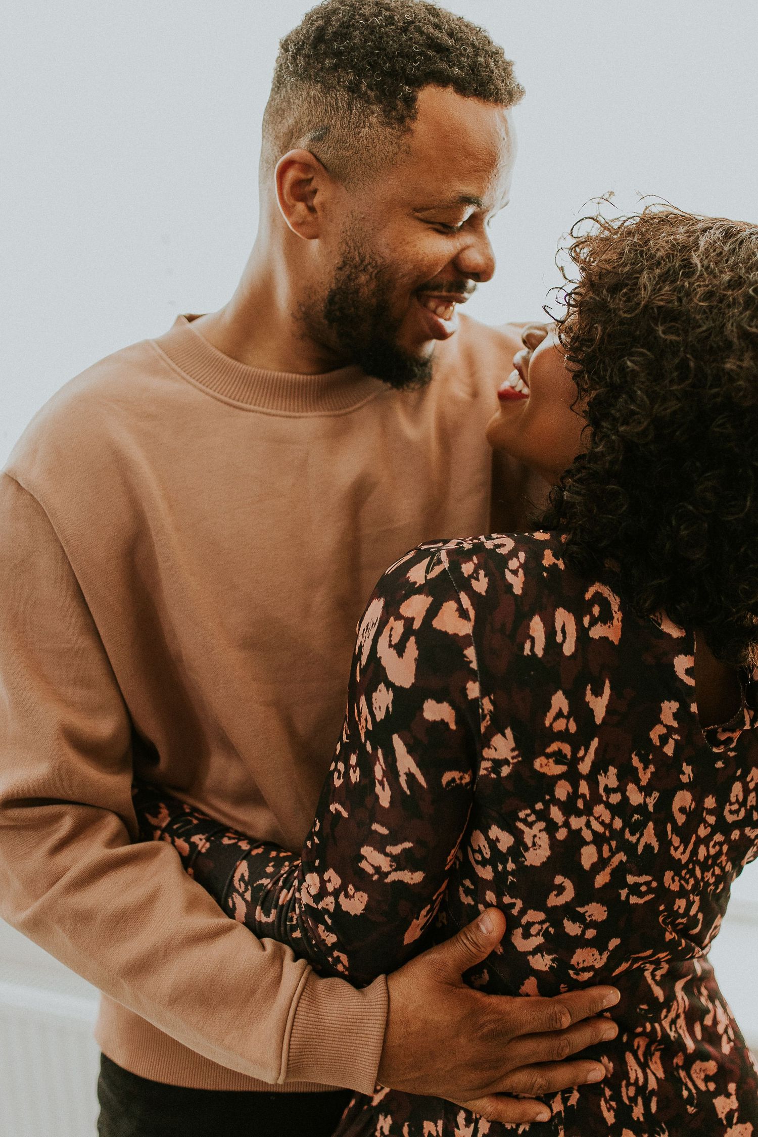 A loving couple shares a tender moment while embracing, wearing a tan sweater and patterned dress against a light background.
