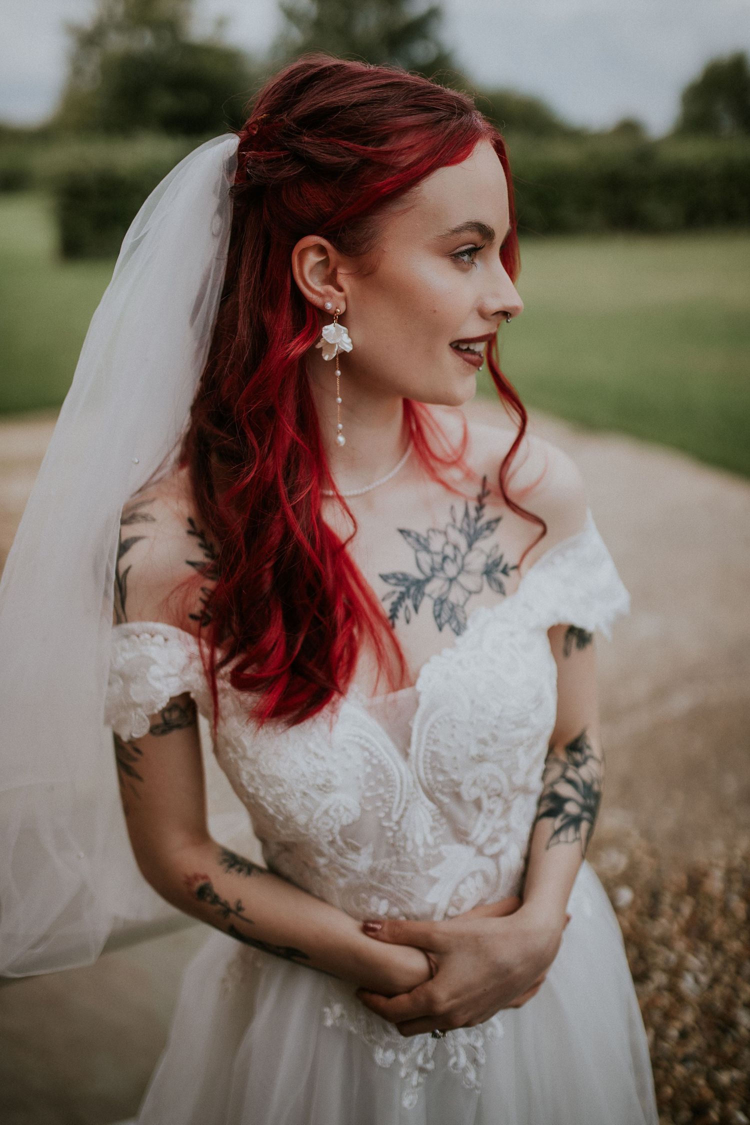 A bride in a lace wedding dress with vibrant red hair and visible tattoos poses in profile outdoors.
