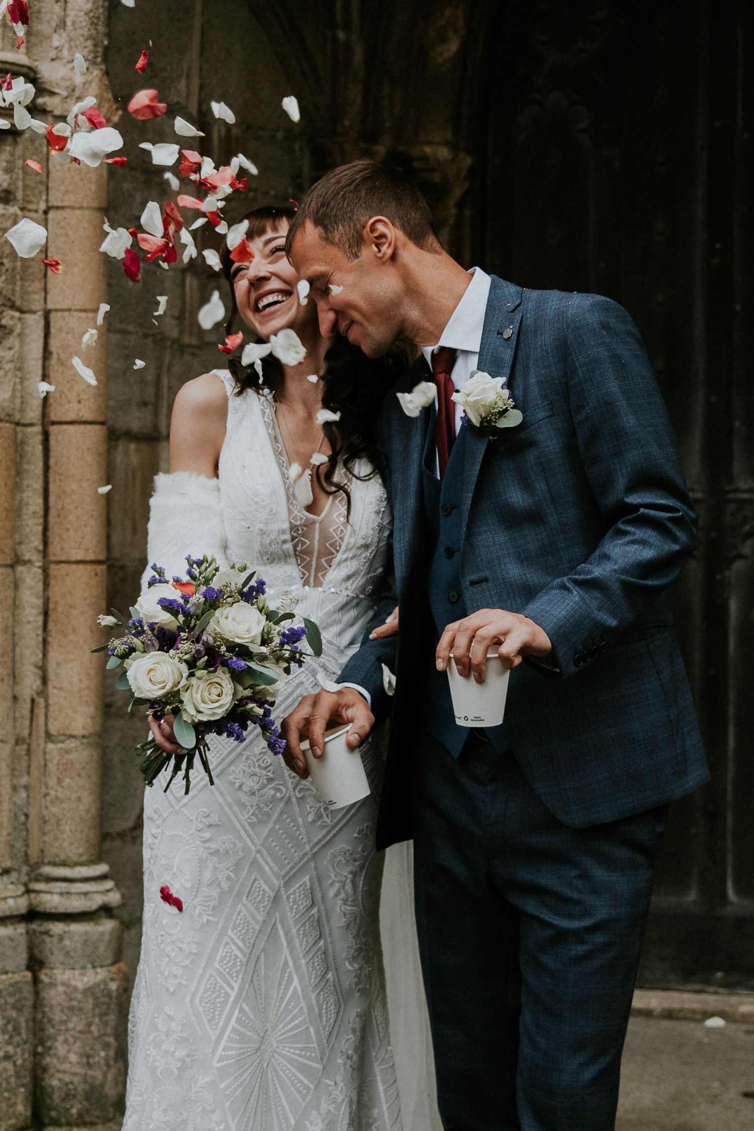 Happy wedding couple laughing while surrounded by flying red and white rose petals.