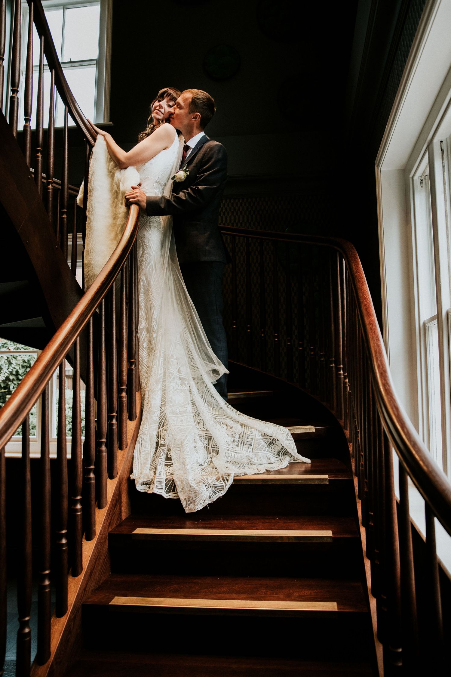 Romantic wedding couple embrace on wooden staircase with natural window light.