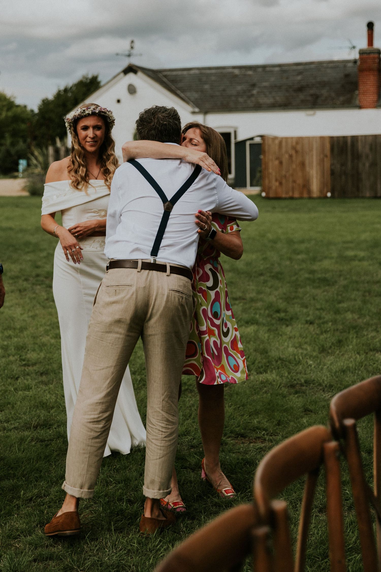 Wedding couple and guest share emotional embrace on grassy lawn with white farmhouse in background.