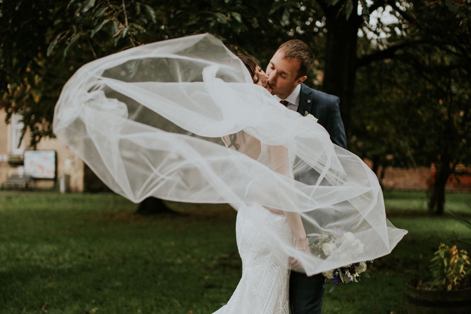 A dramatic wedding photo with flowing white veil billowing in the breeze against a garden backdrop.
