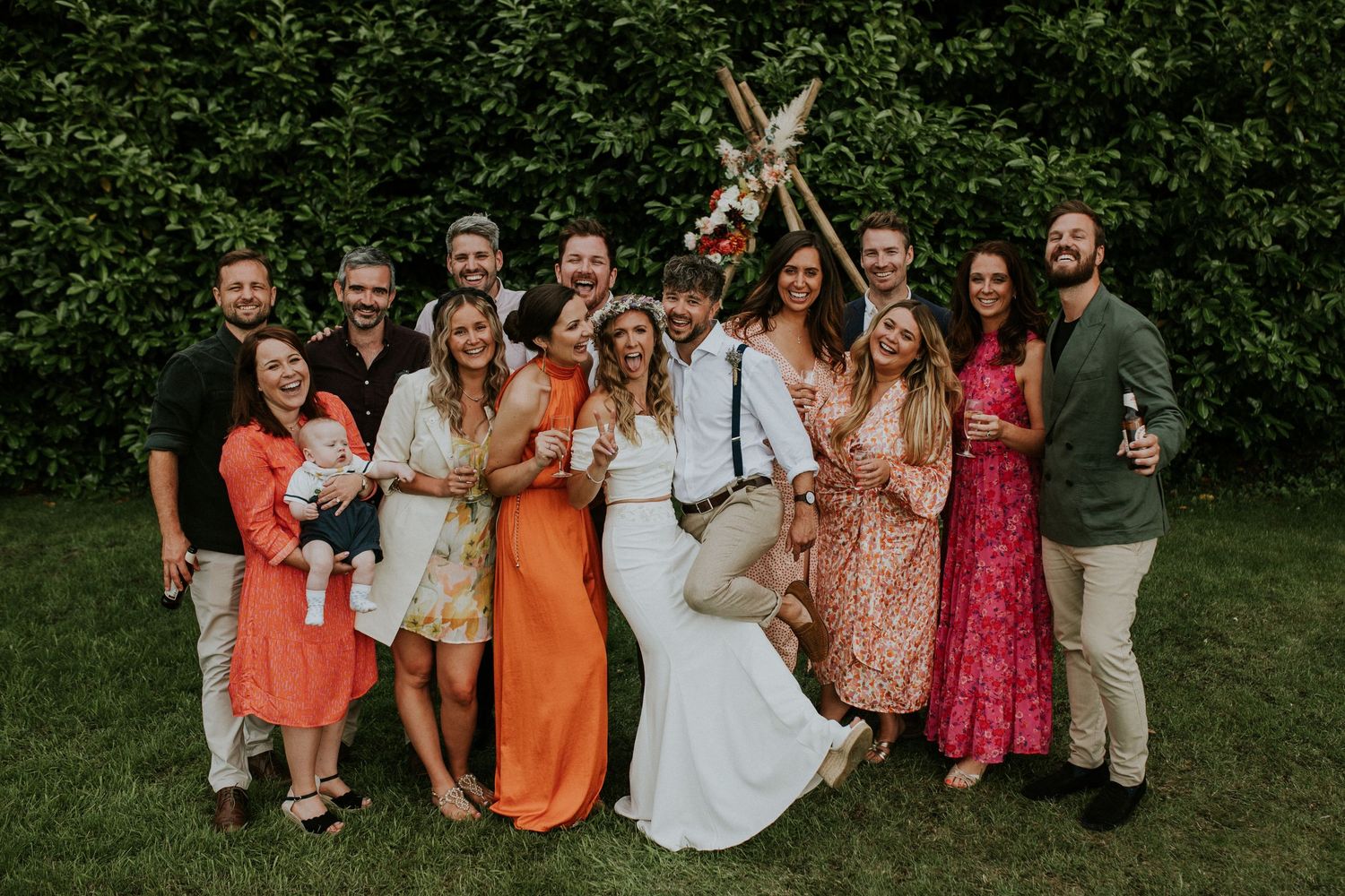 Wedding party poses for group photo in colorful attire during outdoor garden celebration.