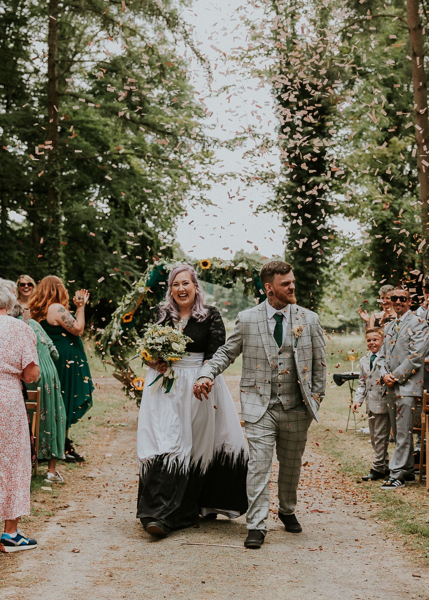 Wedding guests gather outdoors in a mix of formal and casual attire under tree-lined path.