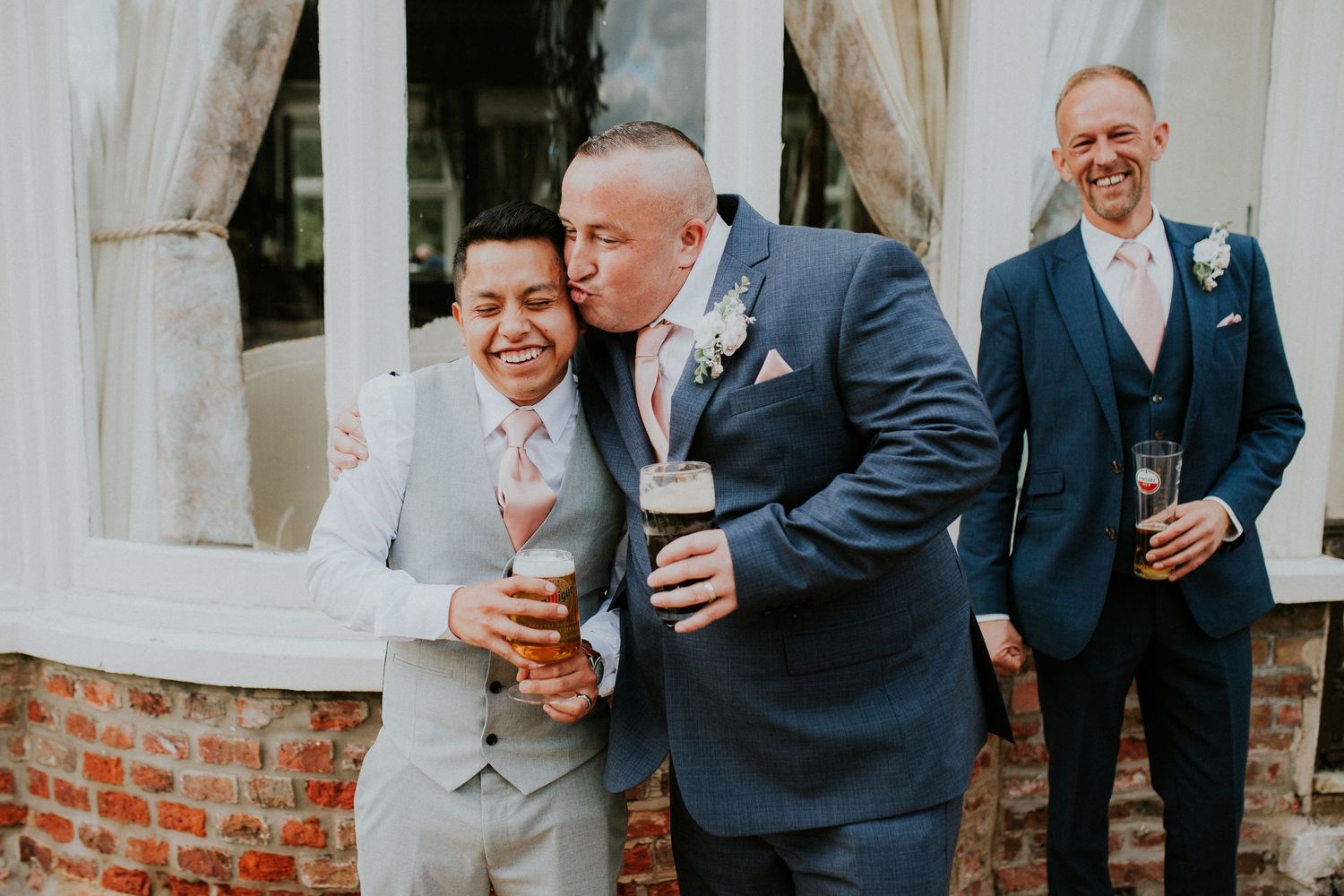 Candid moment of wedding guests sharing drinks and laughter by a brick wall gazebo.