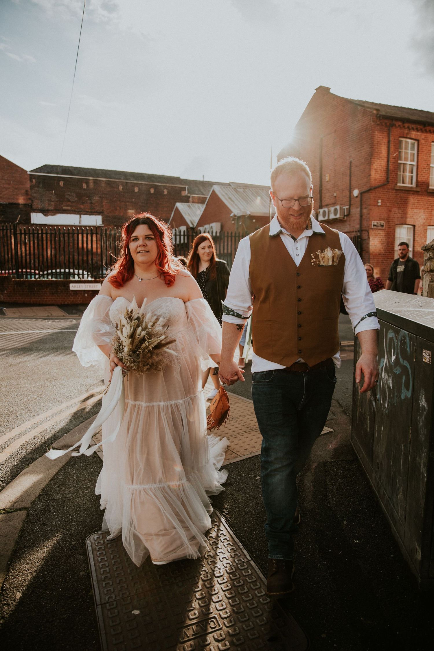 Wedding party walking together on cobblestone street at sunset near brick buildings.