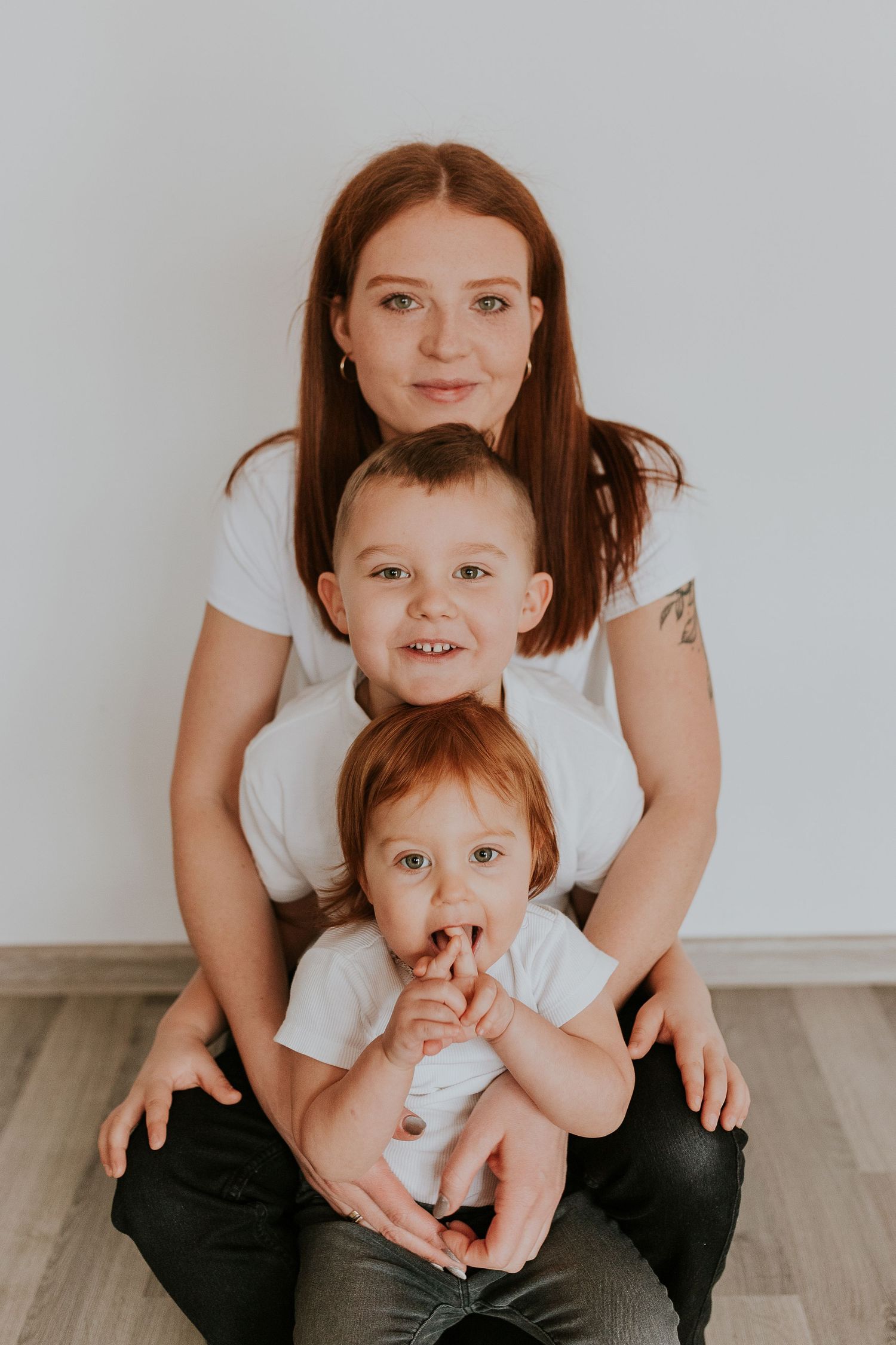 Mother and baby share playful bonding moments on a light wooden floor in matching white outfits.