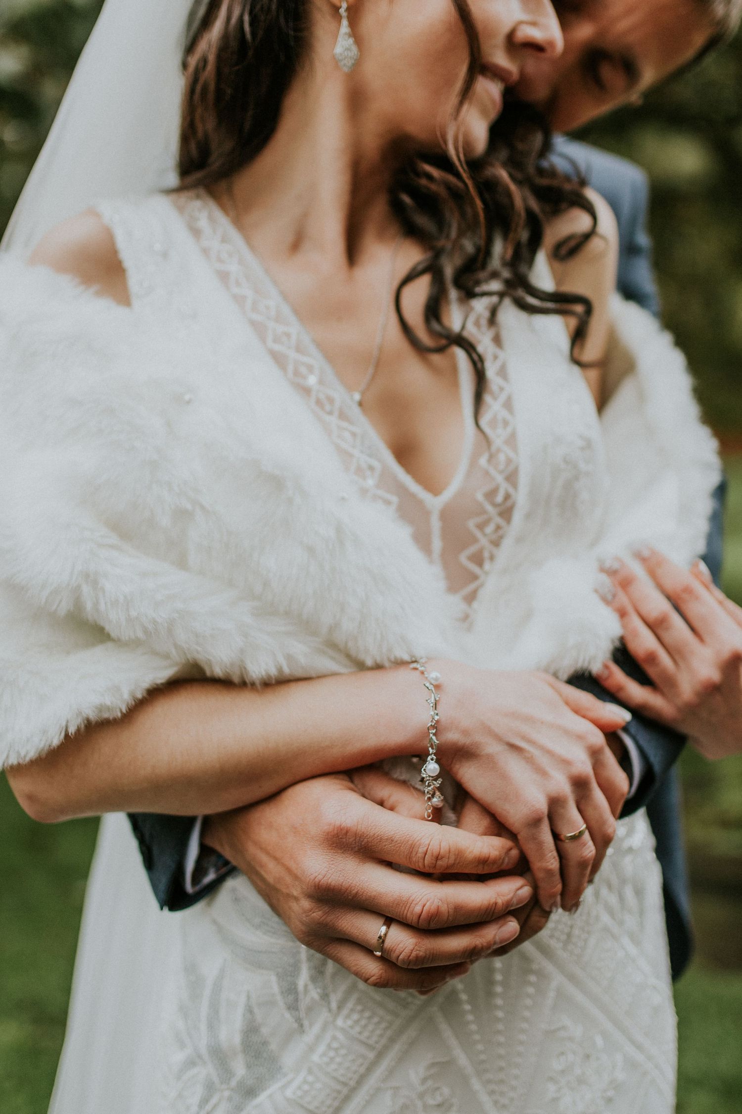 Close-up detail of bride's lace dress and fur wrap with groom's hands embracing from behind.