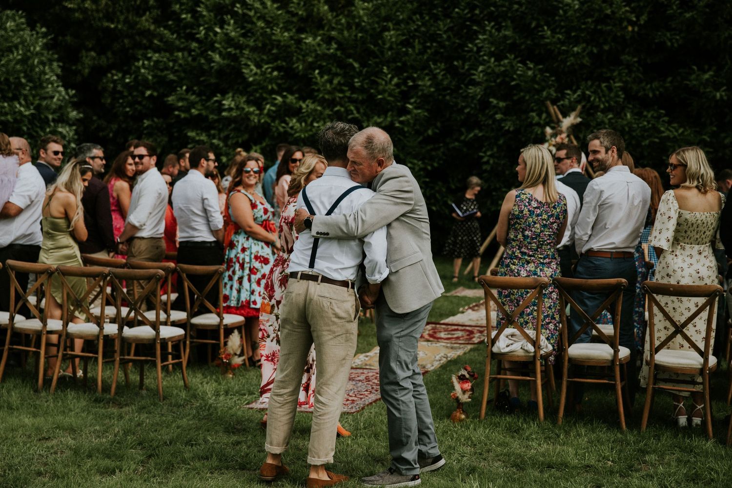 Wedding guests gather on lawn with wooden chairs as couple embraces during outdoor ceremony.