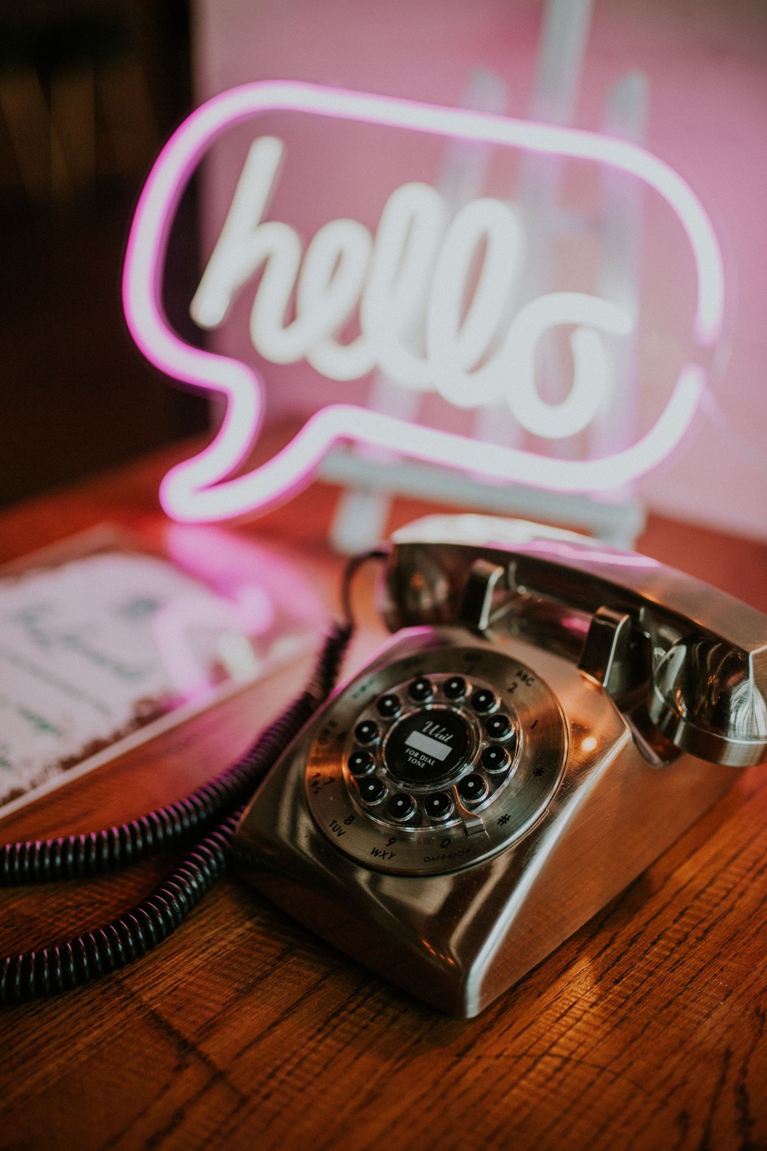 A vintage rotary telephone sits on a wooden table beneath a pink neon 'hello' speech bubble sign.