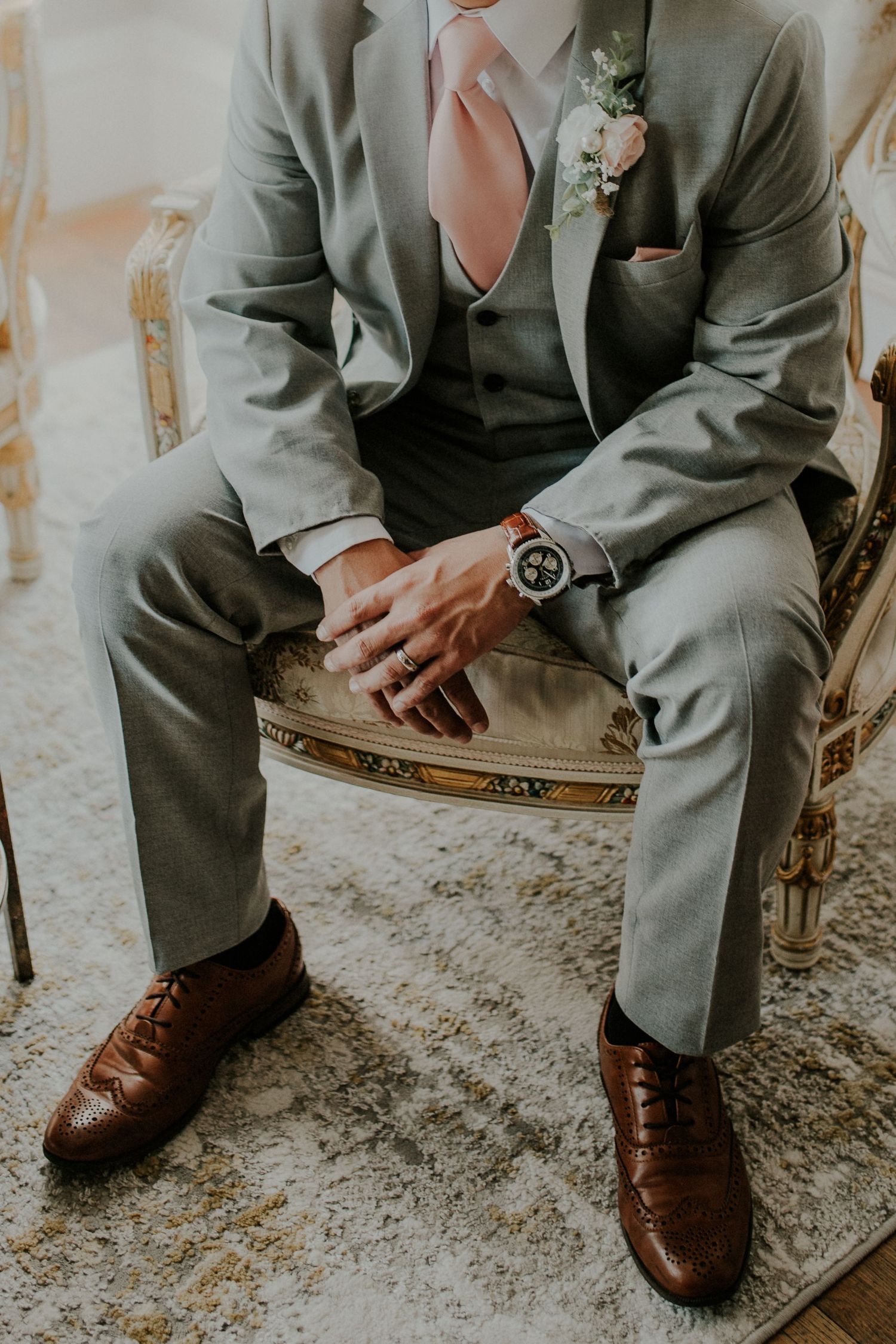 Groom in light grey suit with peach tie and brown oxford shoes sitting in vintage chair.