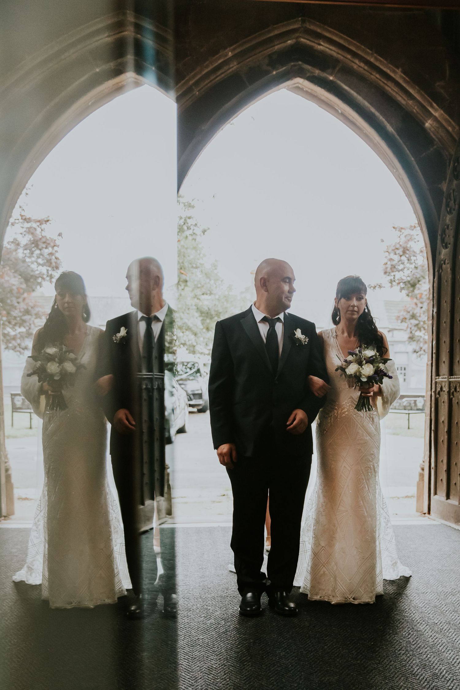 Silhouettes of wedding party members stand beneath historic church archway.