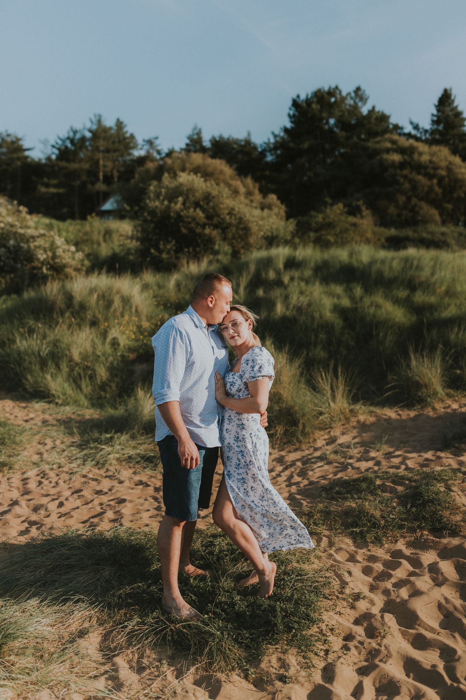 Romantic couple embraces on sandy beach near coastal dunes during golden hour.