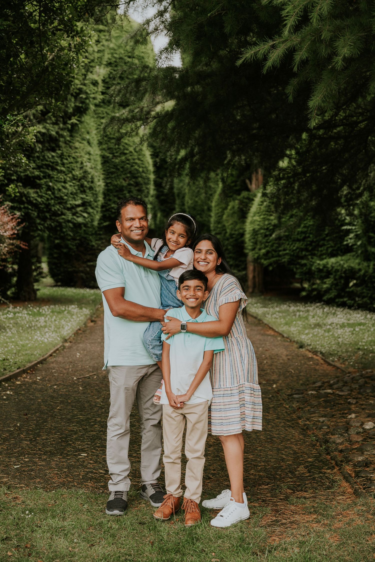 Family wearing coordinated light colored outfits poses on tree-lined path.