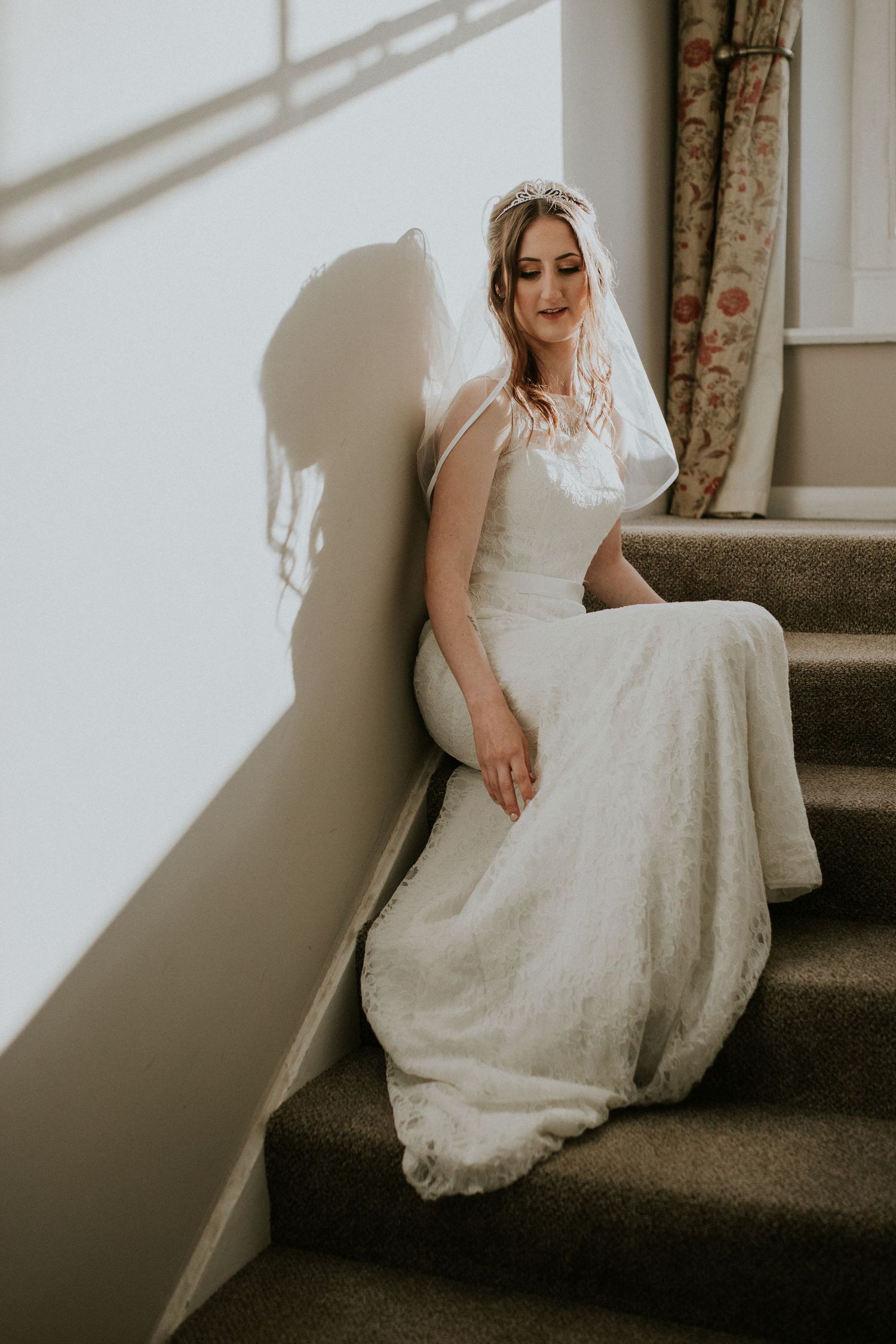 Soft window light illuminates bride's white lace gown on carpeted stairs.