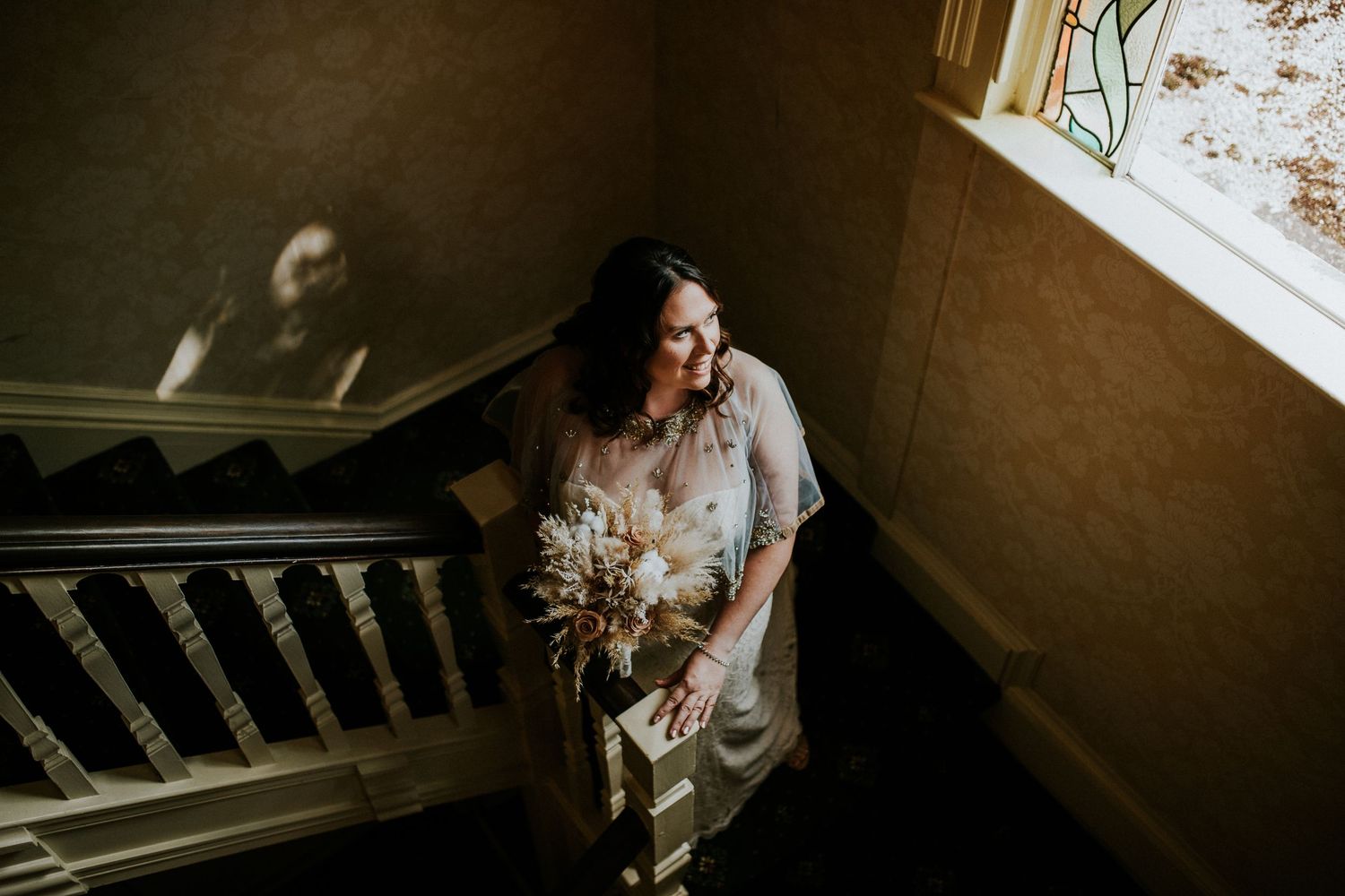 A person in wedding attire pauses on a vintage staircase near a window.