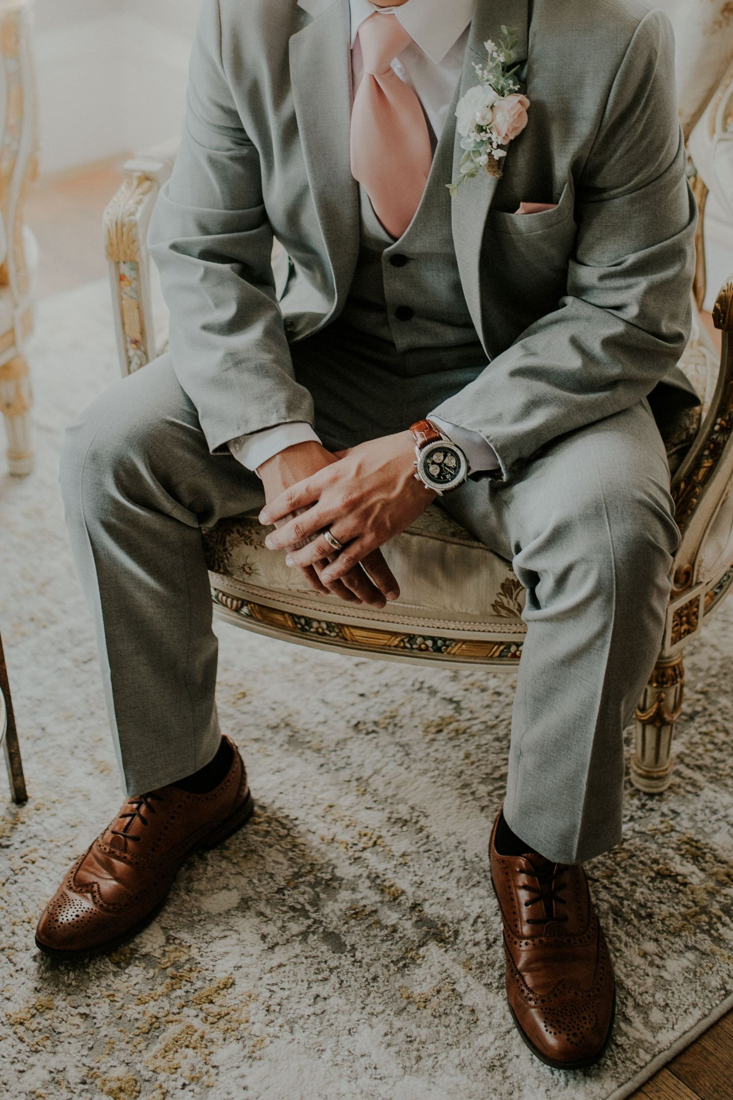 Detail shot of a groom in a gray suit with pink tie and brown leather shoes sitting on an antique chair.