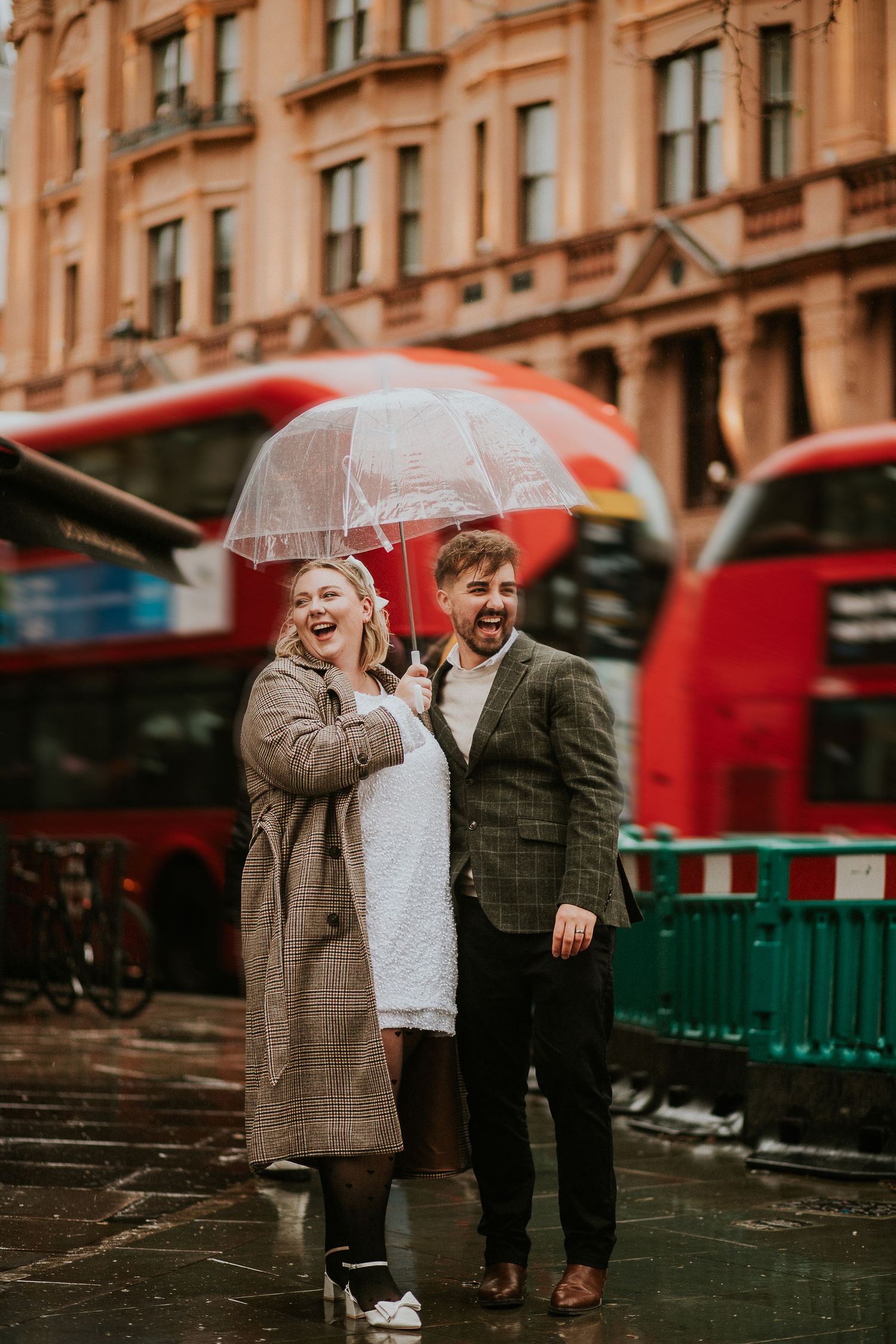 A couple sharing a laugh under a red umbrella on a rainy London street with red buses in the background.