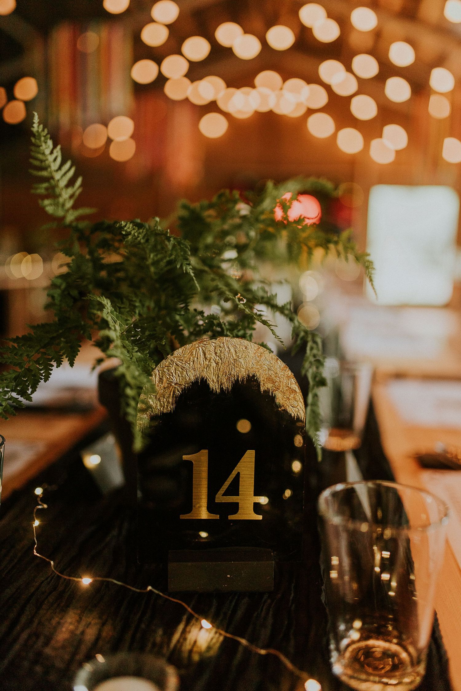 Wedding table decor with golden number 14 sign and greenery against bokeh lights.