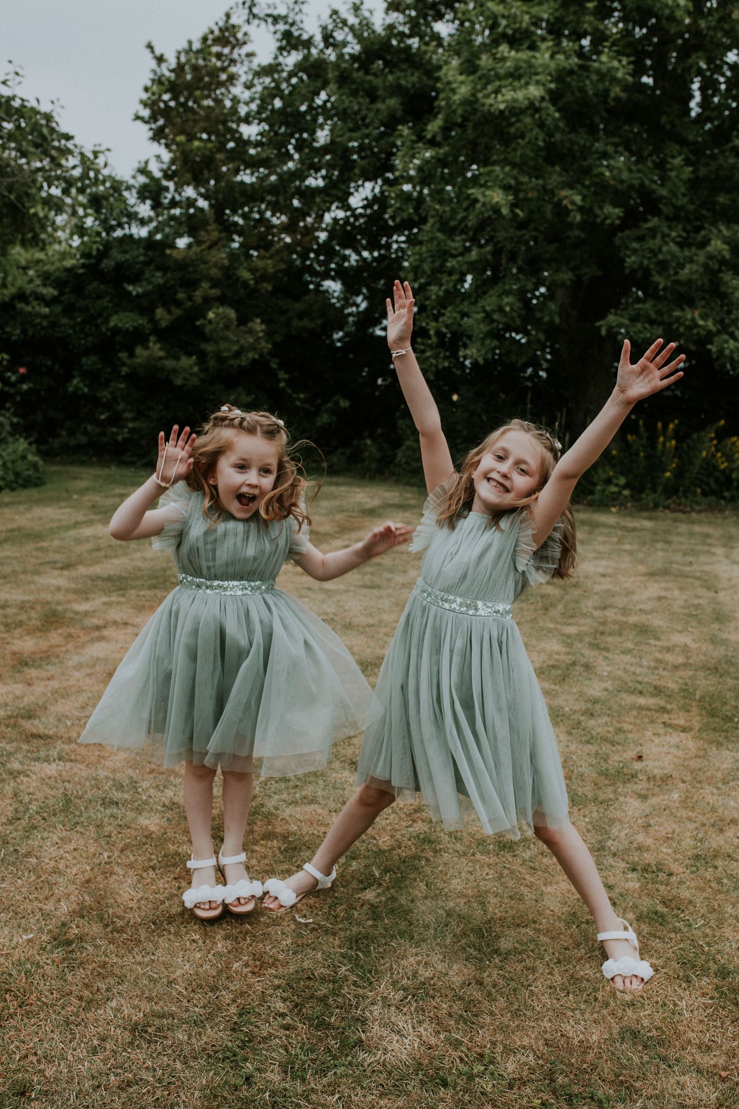 Two young flower girls in sage green dresses jump playfully on grass.