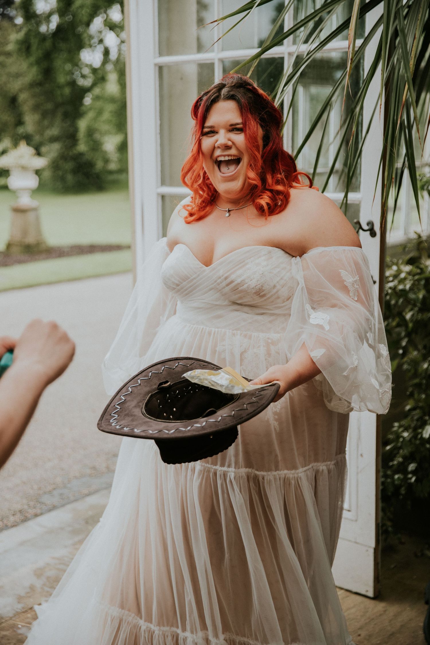 A joyful bride in a flowing white dress laughs while holding a dark hat during an outdoor wedding photoshoot.