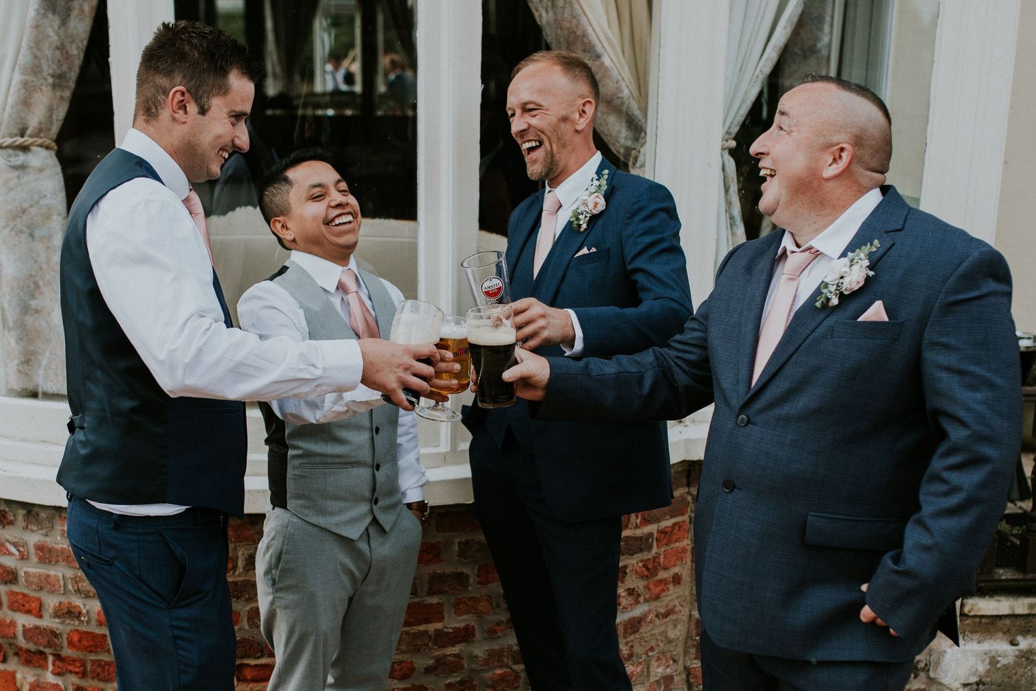 Groomsmen sharing drinks and laughs outside brick venue during wedding celebration.