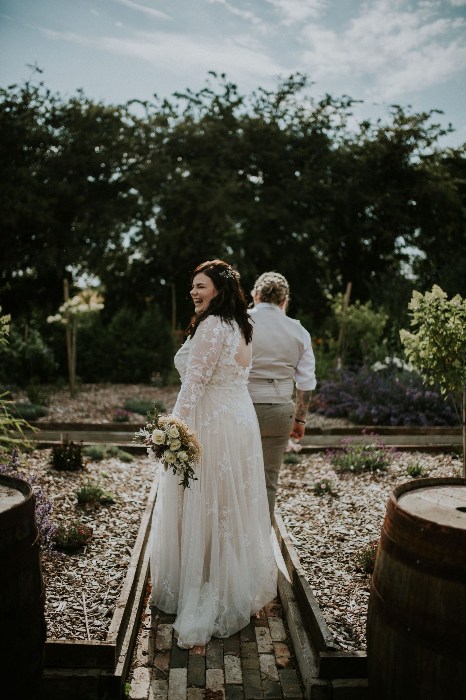A couple stands on a garden path with rustic barrels and greenery in the background.