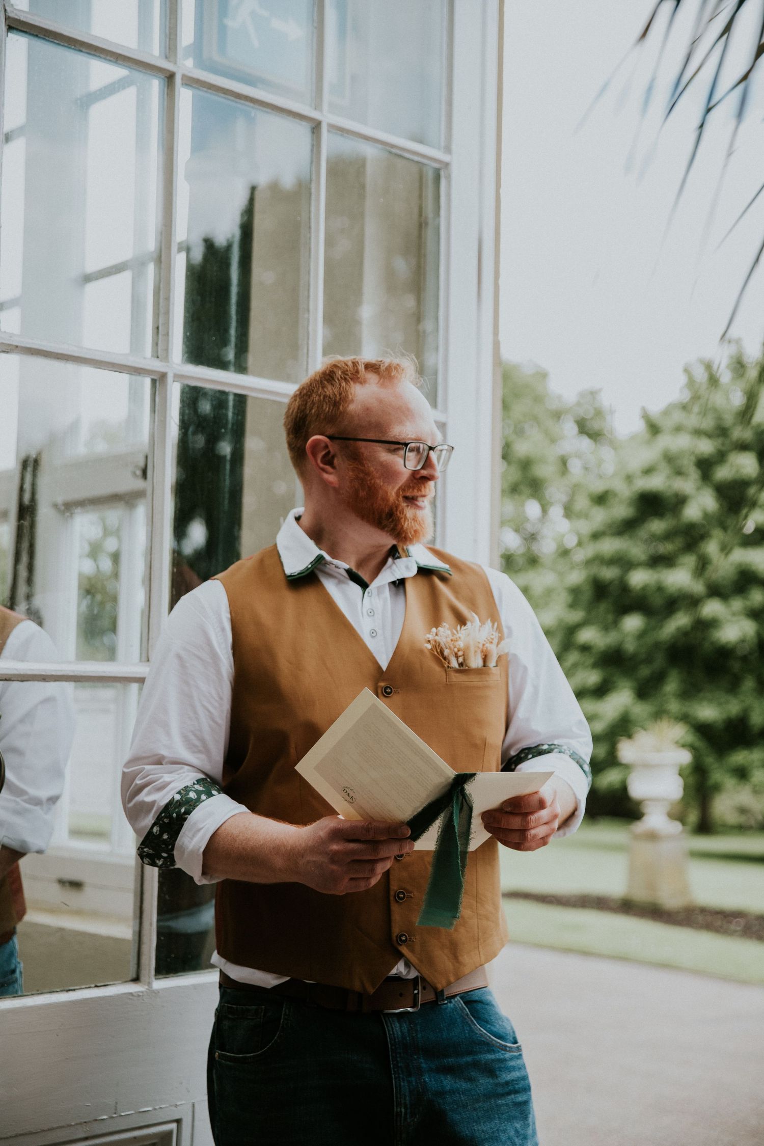 A person in a tan vest reads from papers while standing by large windows.