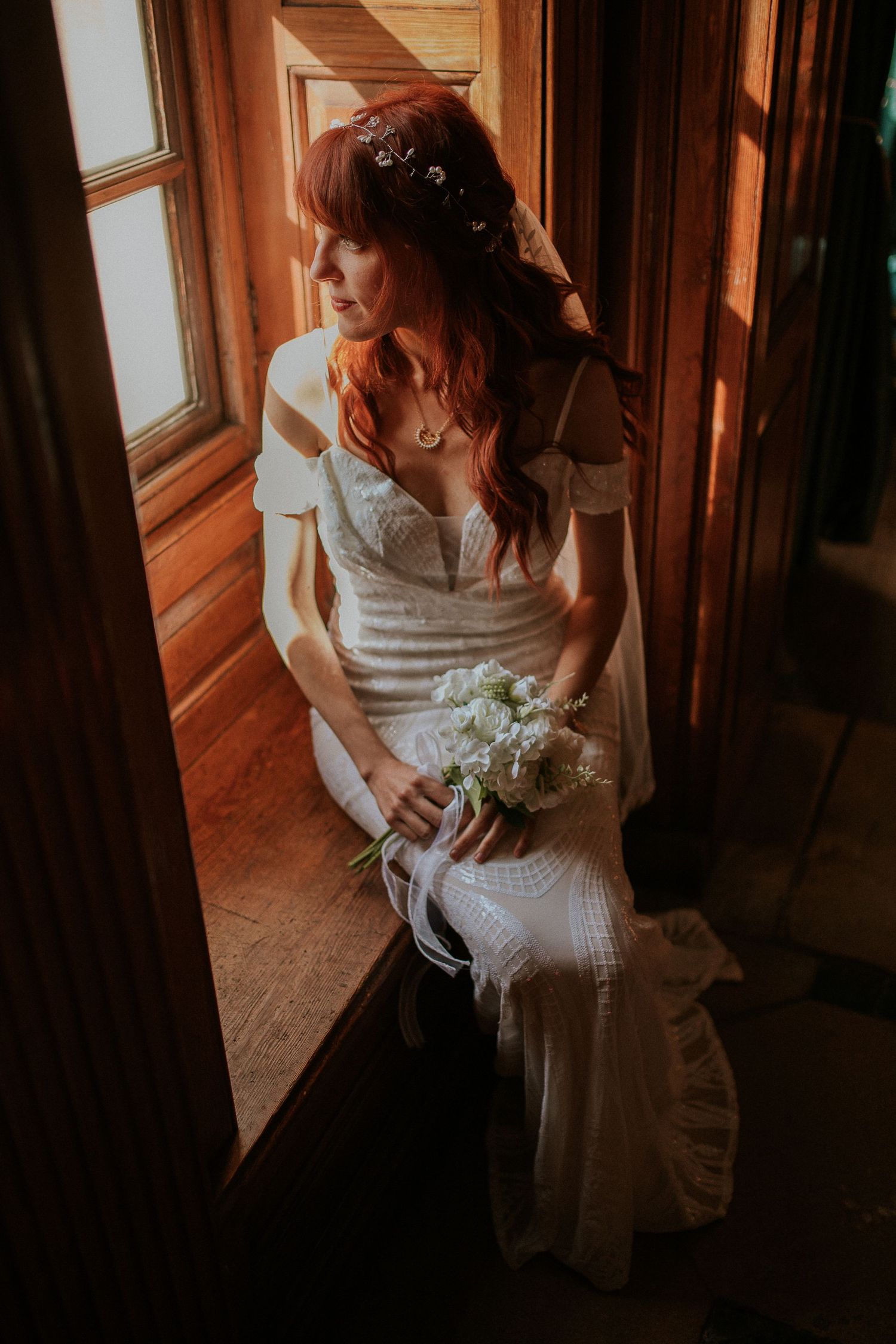 Bride in white lace dress holding bouquet poses near window in warm sunlit room.