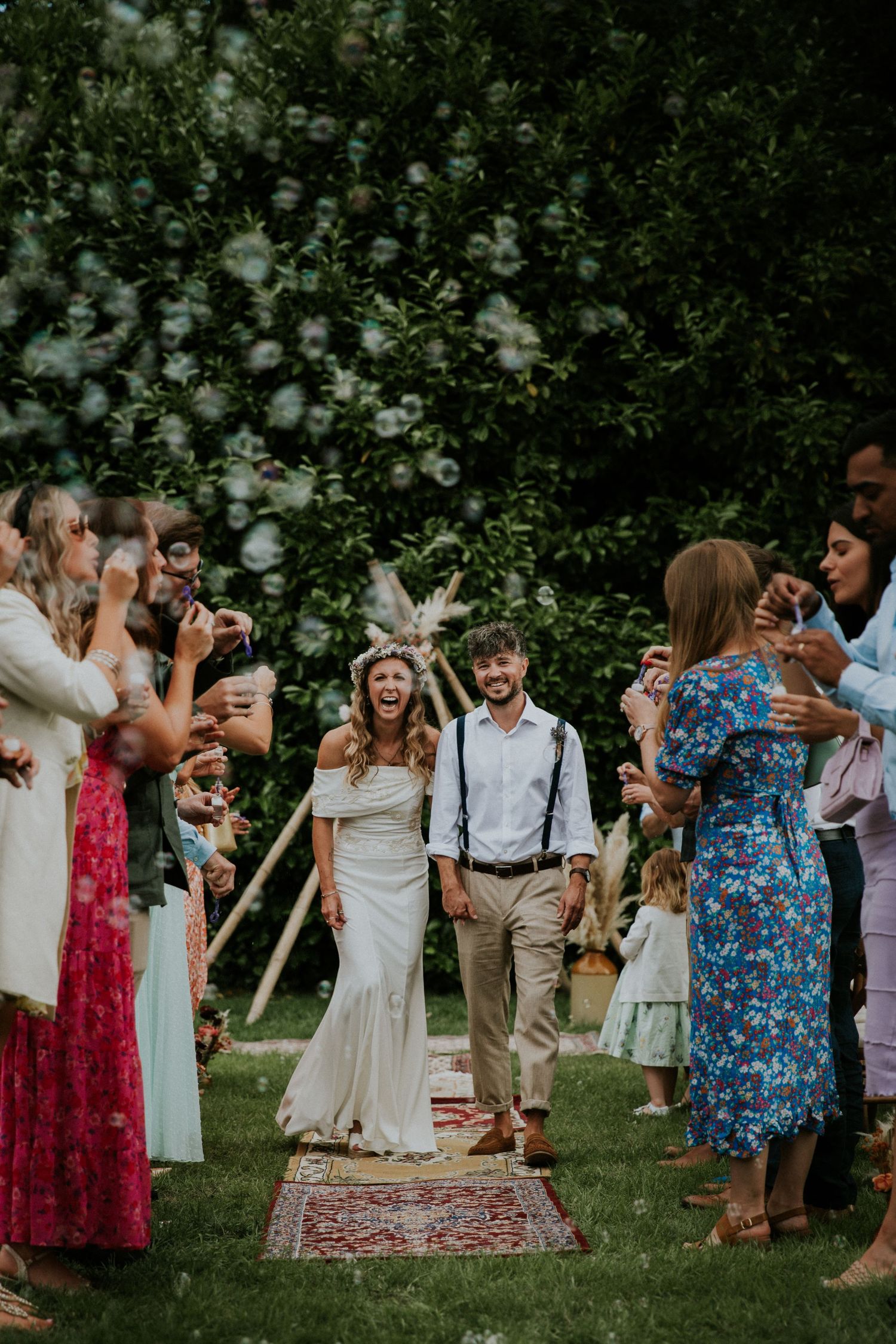 Wedding guests in colorful dresses dancing on a lawn with decorative rugs.