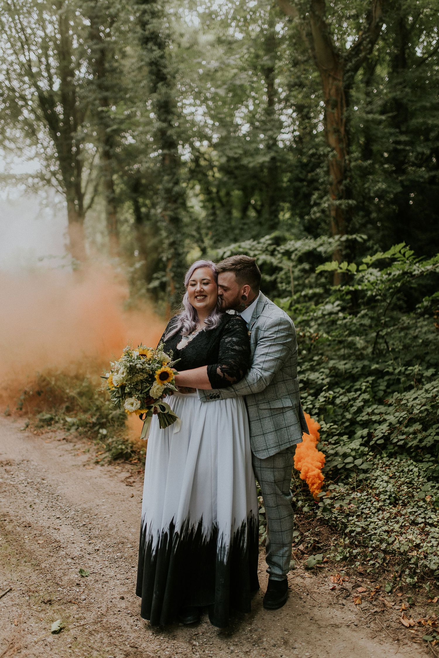 Couple sharing intimate moment with orange smoke bomb on forest path during wedding photoshoot.