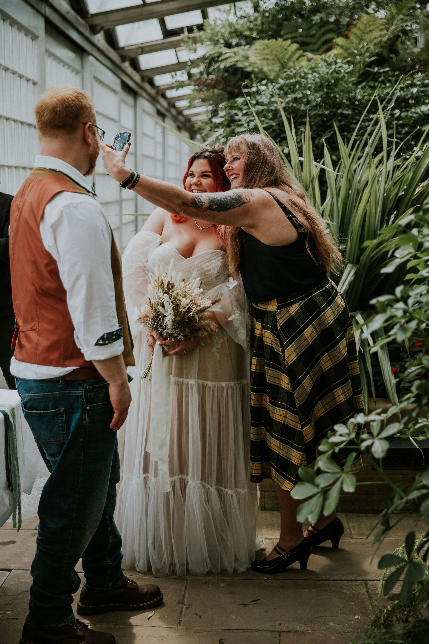 Wedding party taking selfies in a greenhouse setting with natural light streaming through.