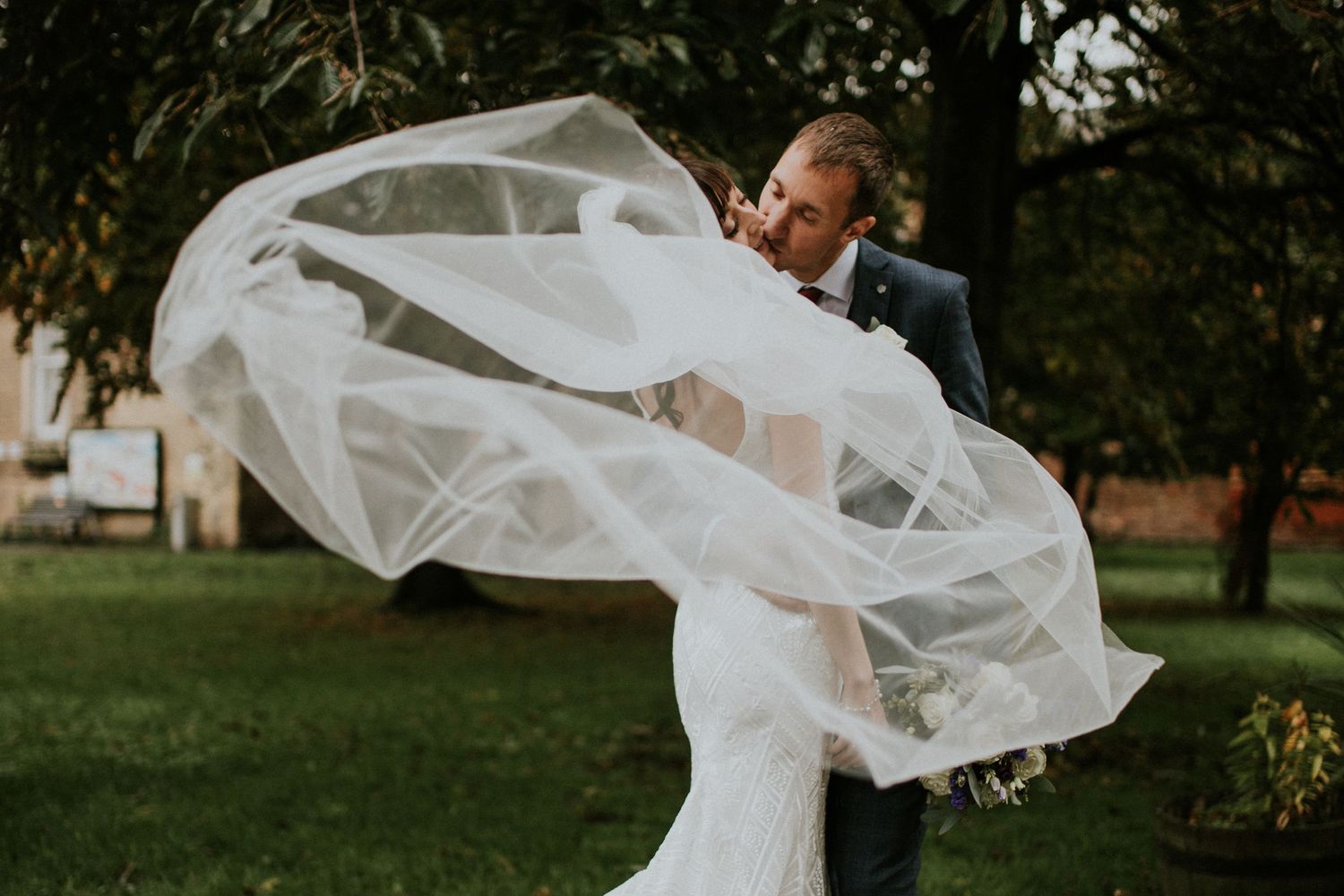 Bride's veil dramatically catches wind as groom watches in garden setting.