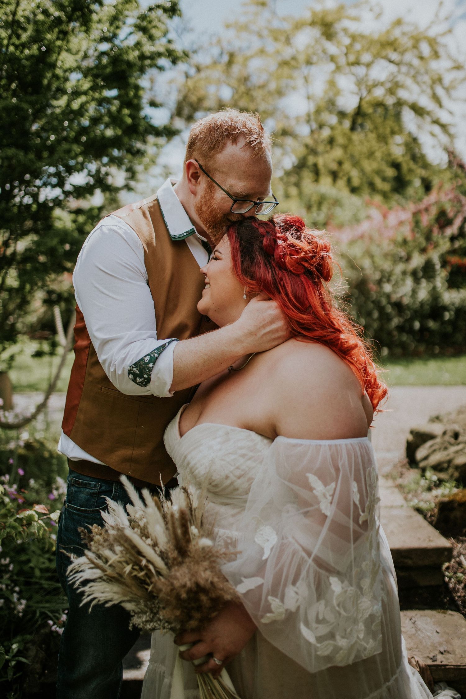 Romantic outdoor wedding portrait with dried pampas grass bouquet and soft sunlight filtering through trees in garden setting.