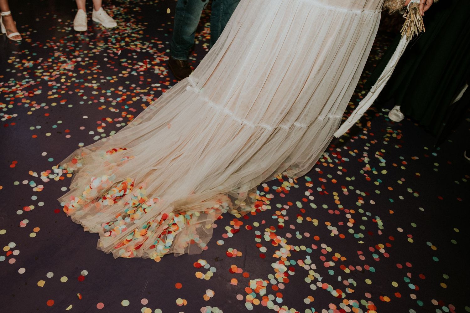 White wedding dress flowing over colorful confetti scattered on dark floor.