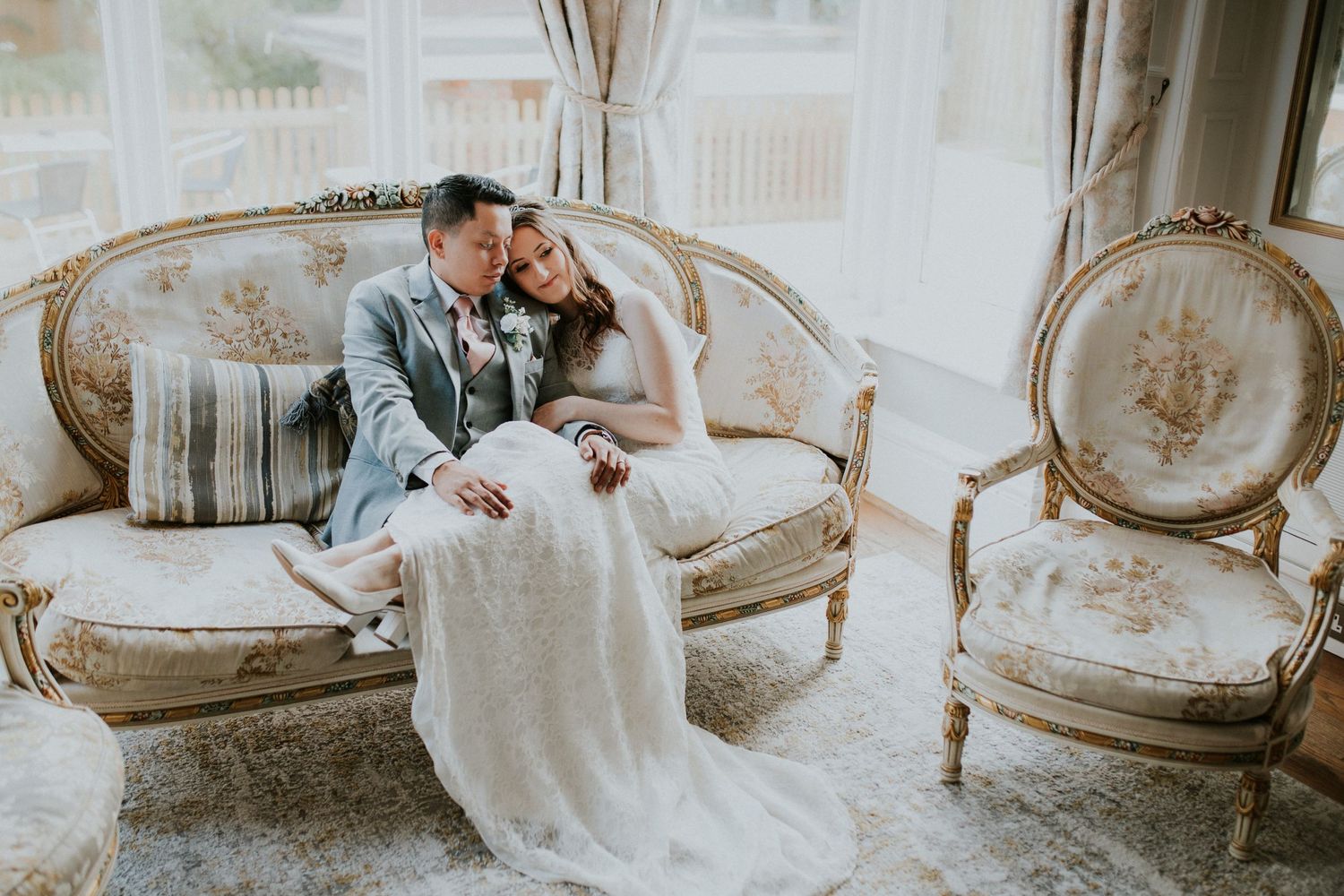 Wedding couple shares intimate moment on vintage French-style settee in sunlit room.