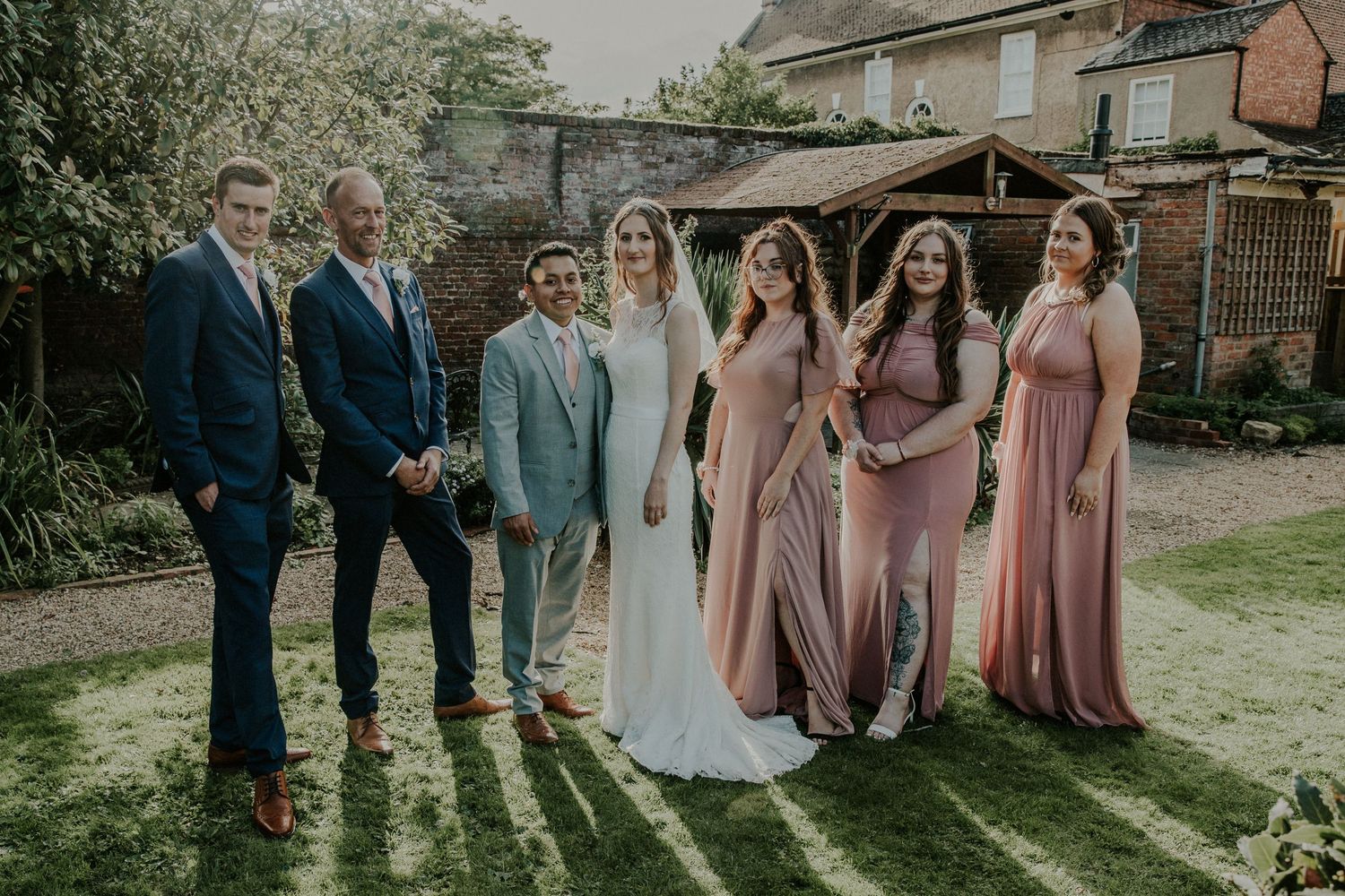 Wedding party in coordinated blue suits and dusty rose dresses poses outdoors on a sunny day.