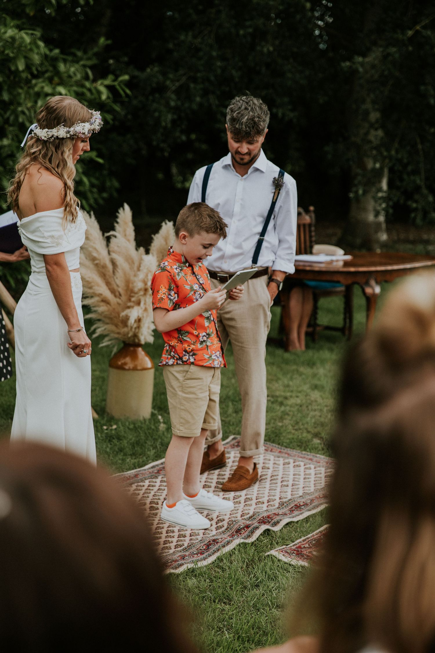 Young attendant reads during outdoor bohemian wedding ceremony with pampas grass decor.