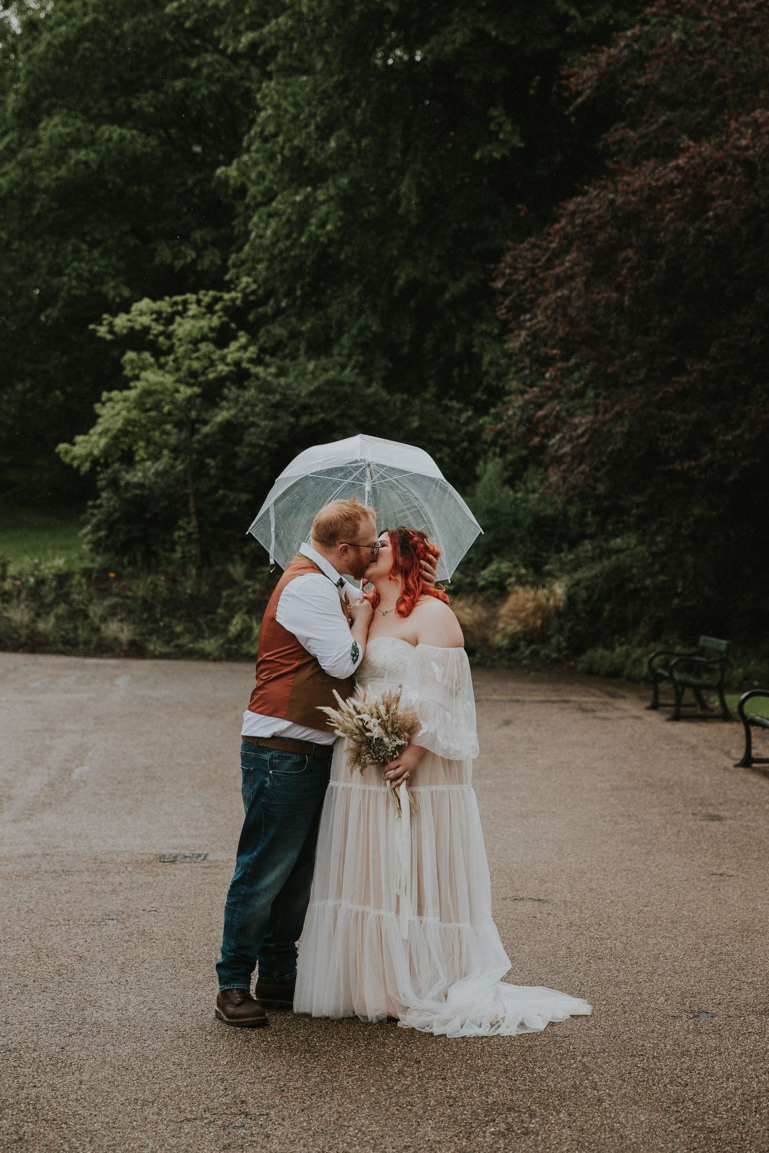 Romantic portrait of newlyweds sharing intimate moment under clear umbrella in garden setting.