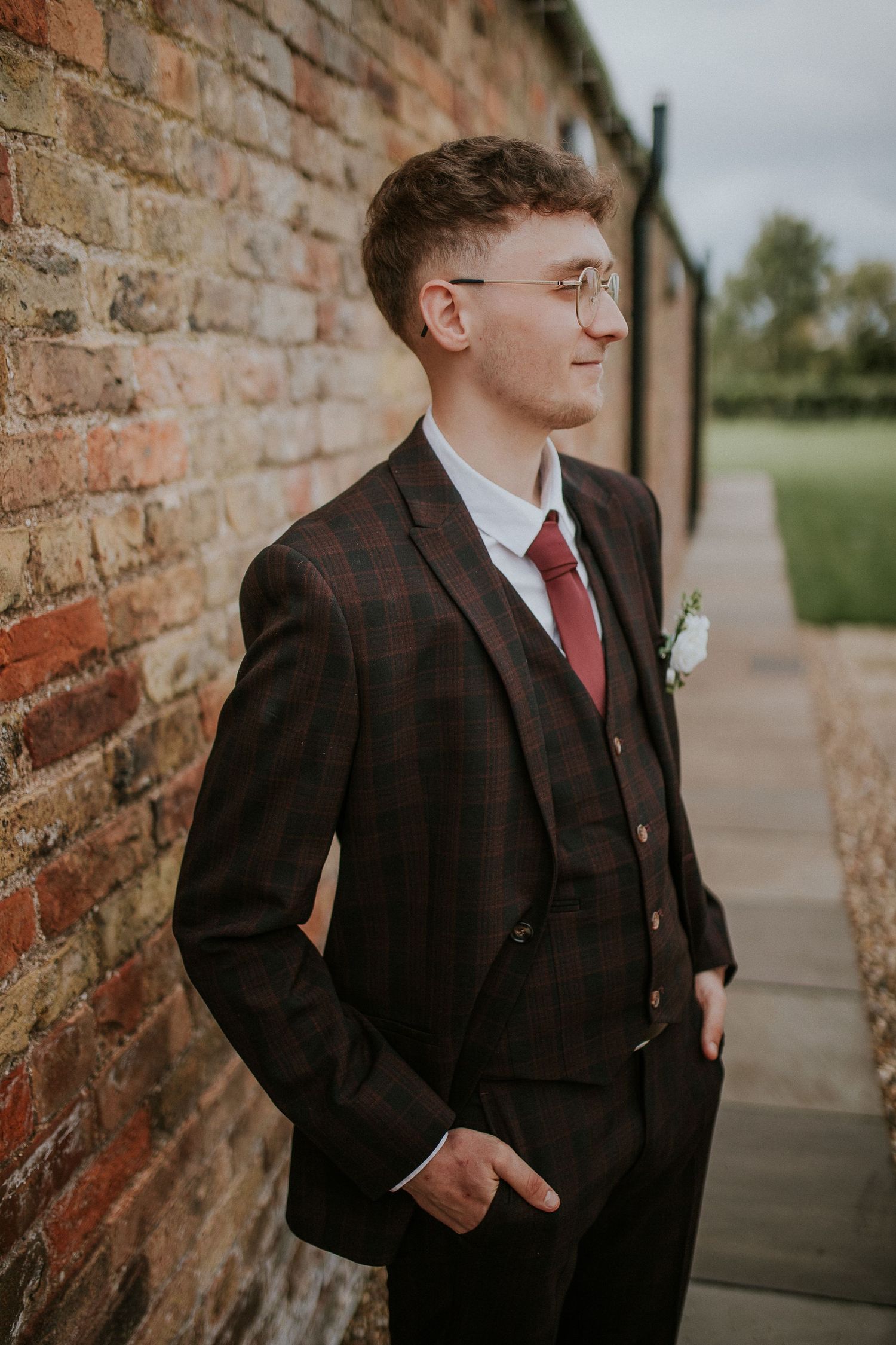 A groom in a plaid suit and round glasses stands against a rustic brick wall.