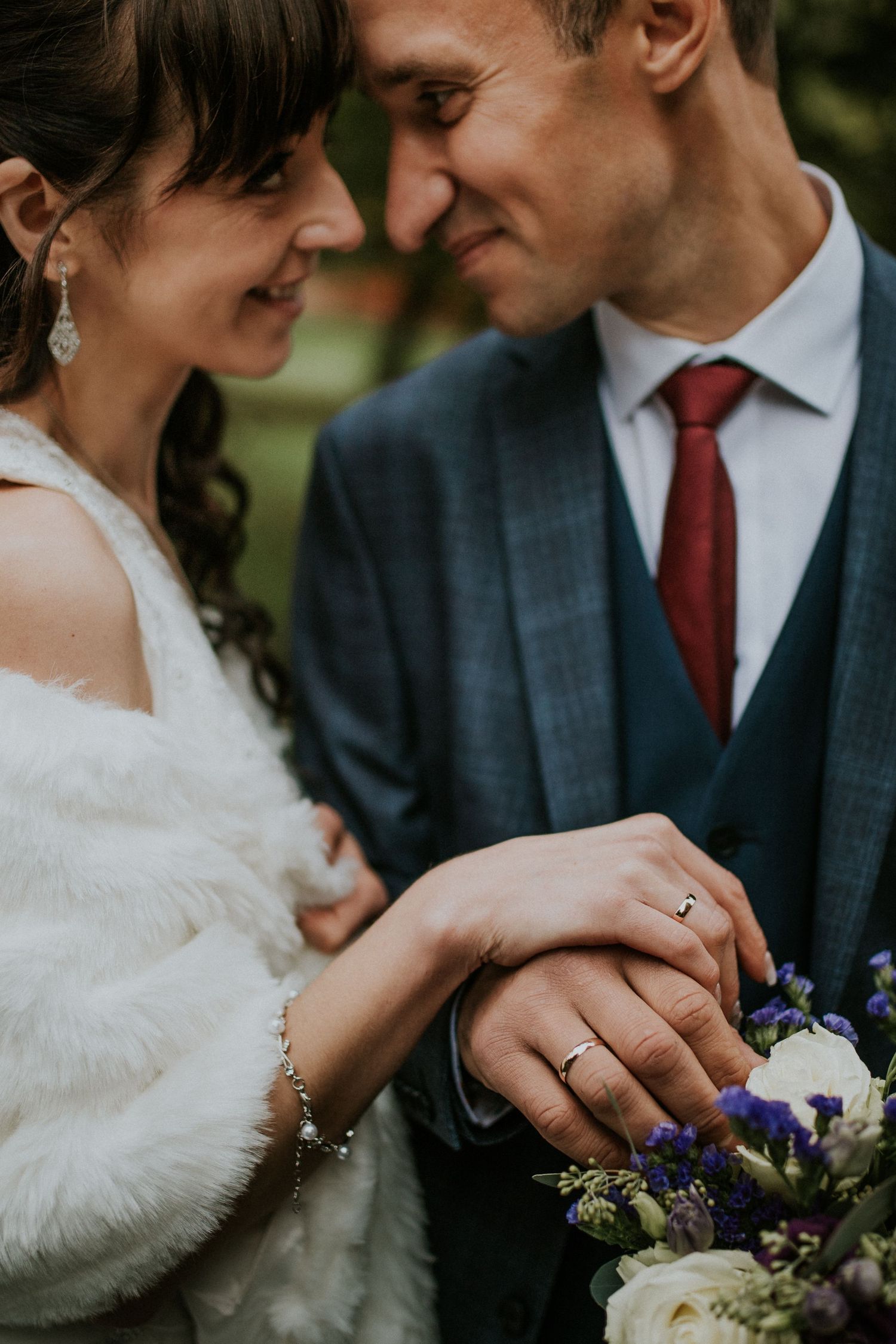 A close-up wedding photo showcasing delicate jewelry and rings against a bouquet of purple flowers.