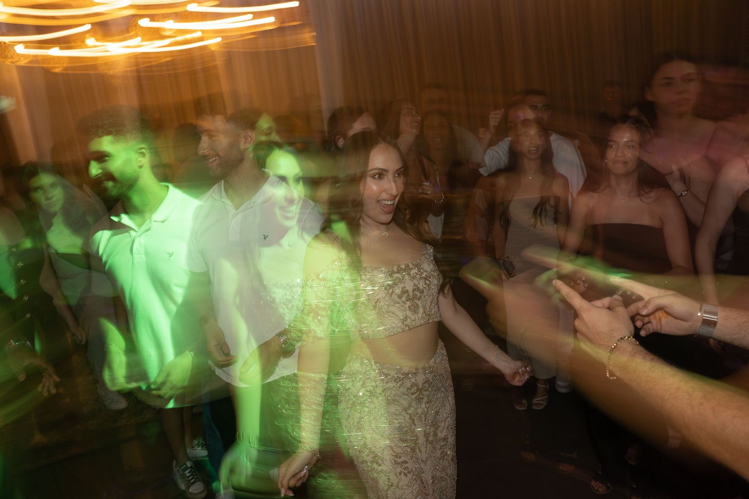 Guests dancing at a glamorous wedding reception under green lights, woman in embellished gown at center.