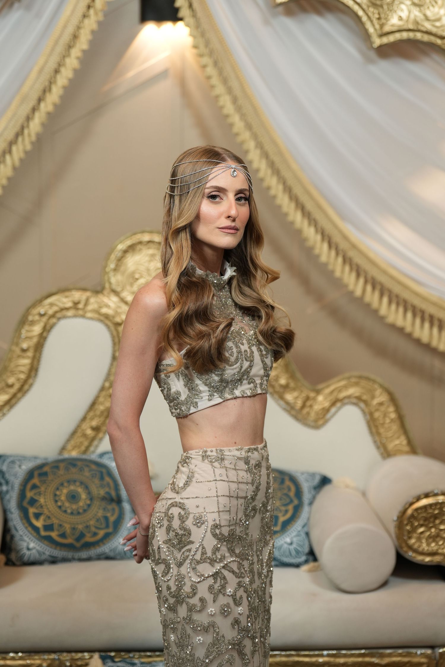 Woman in ornate gold embroidered crop top and skirt with headpiece, posing in elegant gold-trimmed interior.